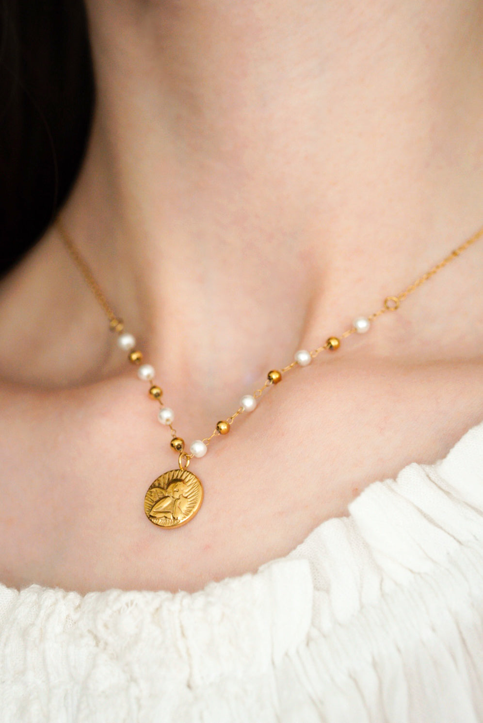 Tuvplāns uz sievietes kakla, uz kuras kakla ir Kaklarota - Amors no Como Jewelry, ar baltām un zelta pērlītēm un apaļu kulonu. Viņa ir tērpusies baltā, teksturētā topā bez plecu malas.