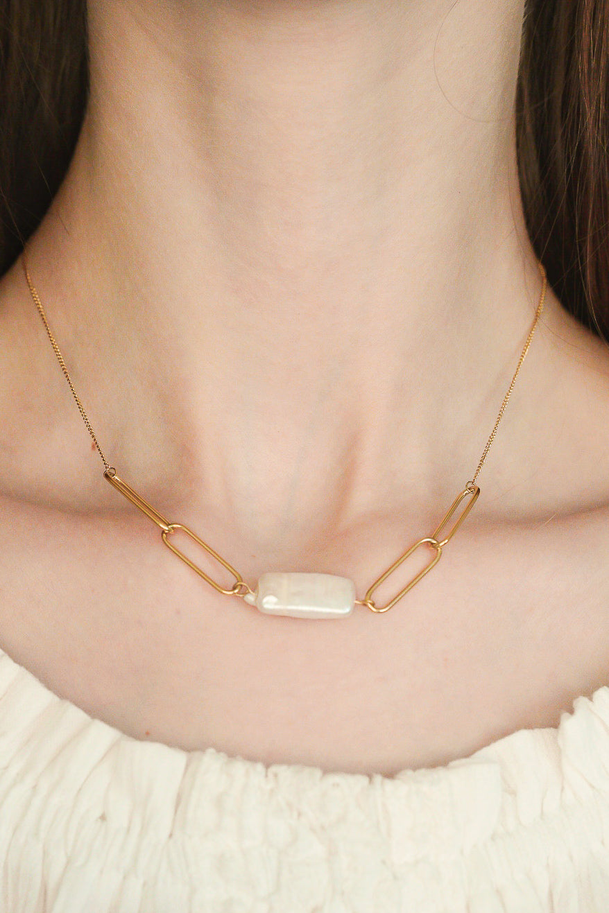 Tuvplānā redzams sievietes kakls, kam mugurā ir eleganta Como Jewelry Kaklarota - Siltums kaklarota ar trim lieliem zelta posmiem un taisnstūra formas balta akmens centrālo daļu, kas rotāta ar baltu rūtainu virsu.