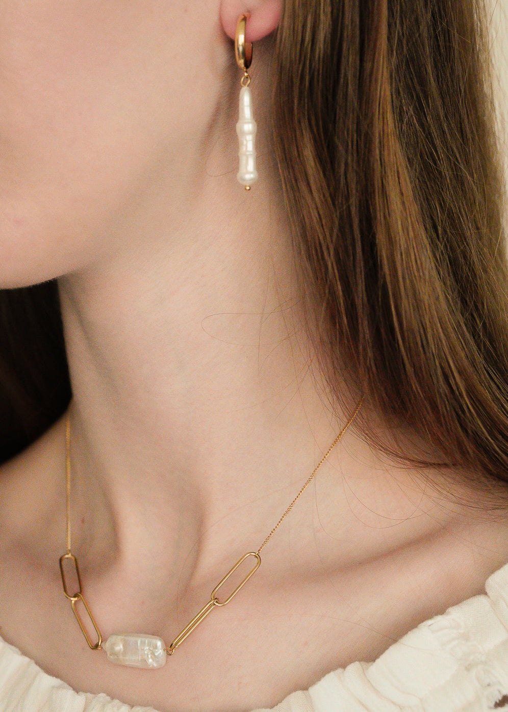 Tuvplānā redzama sieviete krēmīgā topā, kurai ir Como Jewelry Kaklarota - Siltums kaklarota ar taisnstūrveida posmiem un pērļu akcentu, kā arī pieskaņoti zelta auskari ar baltām neregulāras formas pērlēm.