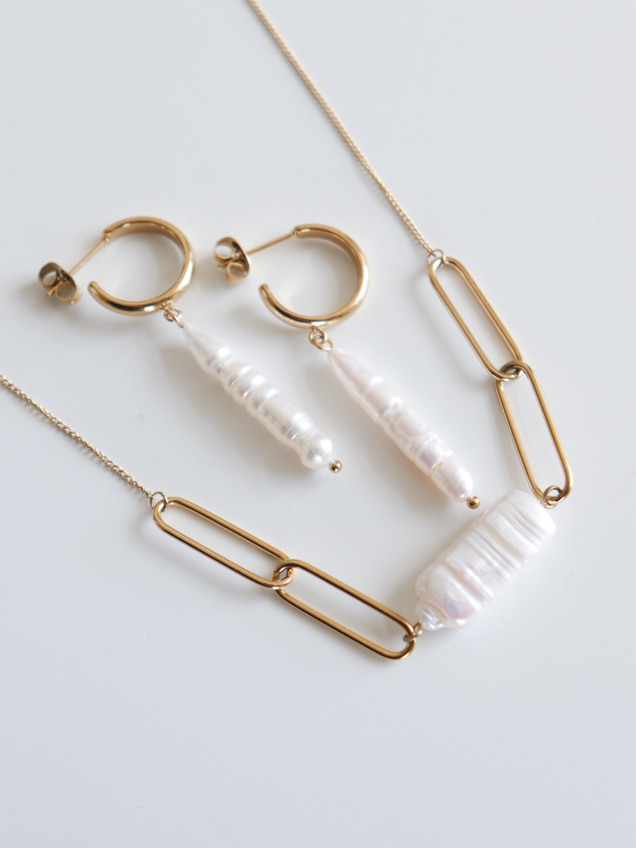 Zelta rotaslietu komplekts uz baltas virsmas, kurā ietilpst divi auskari ar garām, neregulārām baltām pērlēm un Como Jewelry zelta ķēdīte "Kaklarota - Siltums" ar lielu taisnstūrveida baltu pērli.