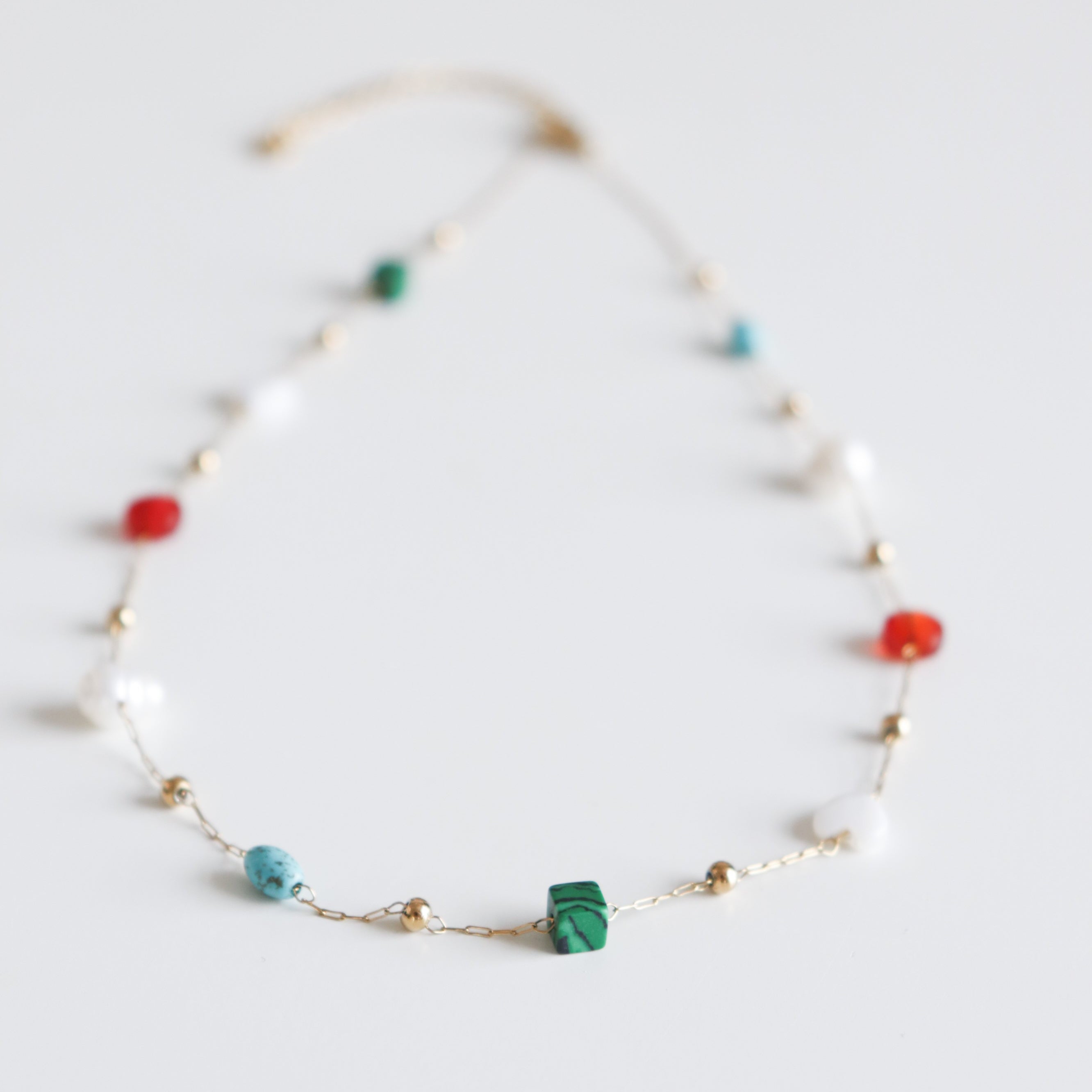 Como Jewelry "Kaklarota - Krāsu rotaļa" ir smalka zelta kaklarota sievietēm ar mazām zaļām, tirkīza, sarkanām un baltām pērlītēm, kas vienmērīgi izvietotas uz zelta ķēdītes uz balta fona.