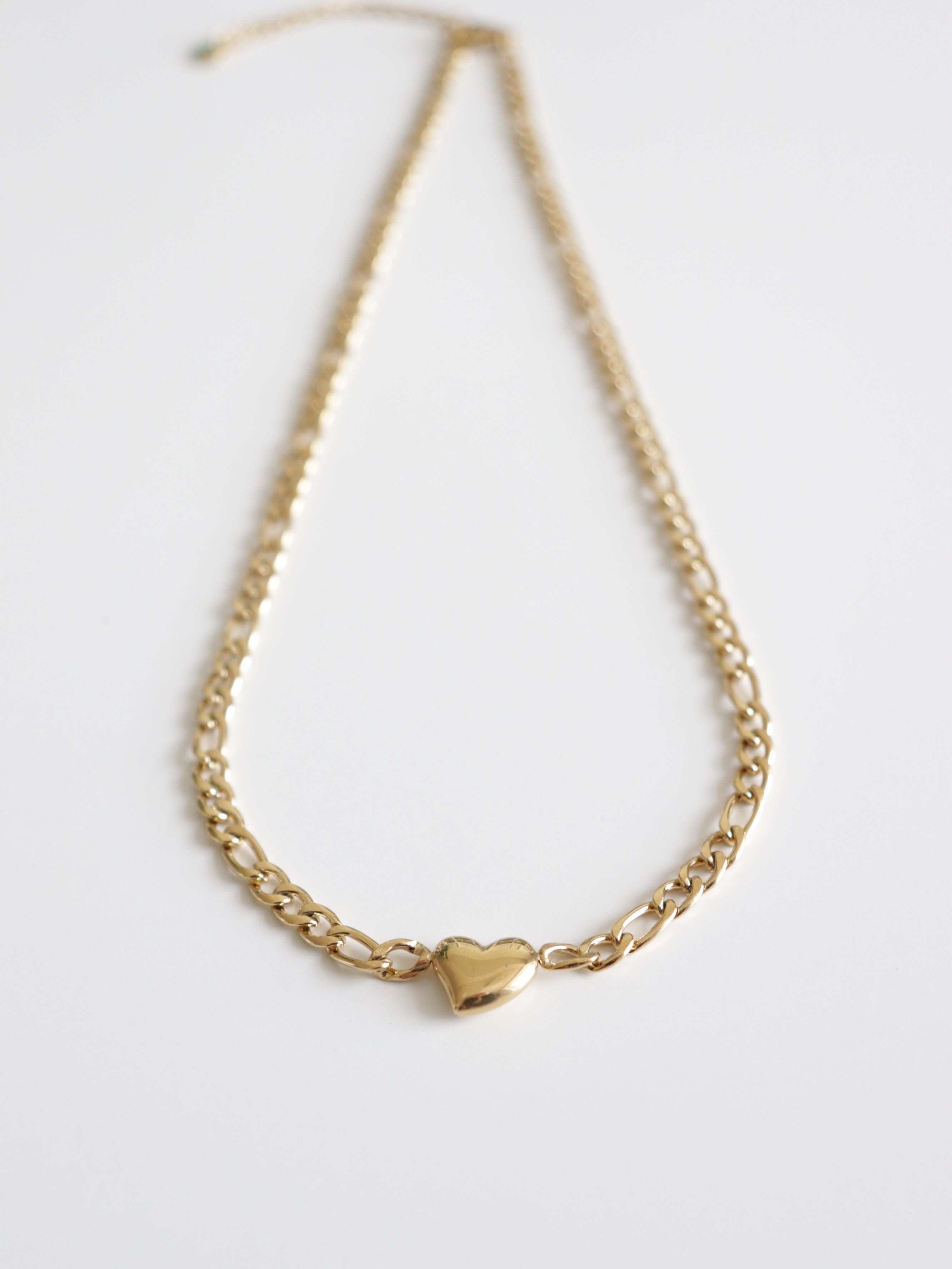 Kaklarota - Sirds no Como Jewelry ar zeltītu ķēdīti un nelielu spožu sirds formas kulonu centrā, attēlota uz balta fona.