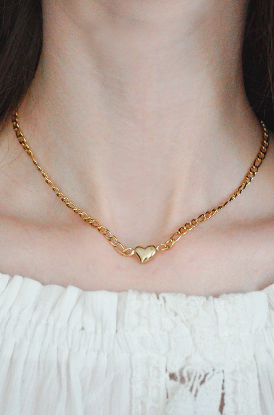 Tuvplānā redzama persona, kas valkā Kaklarota - Sirds by Como Jewelry, modernu zelta ķēdītes kaklarotu ar mazu sirds piekariņu, kas ir kombinēta ar baltas krāsas faktūru.