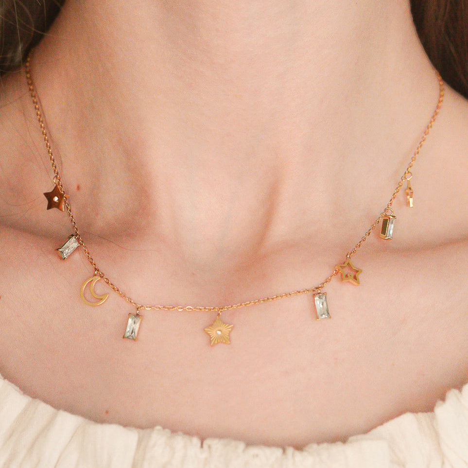 Tuvplāns uz sievietes kakla, kurai mugurā ir Como Jewelry Kaklarota - Zvaigžņu deja, smalka zelta kaklarota ar zvaigznēm, mēnesi un kristāla piekariņiem. Neitrāls fons izceļ viņas krēmkrāsas topu bez plecu malas.