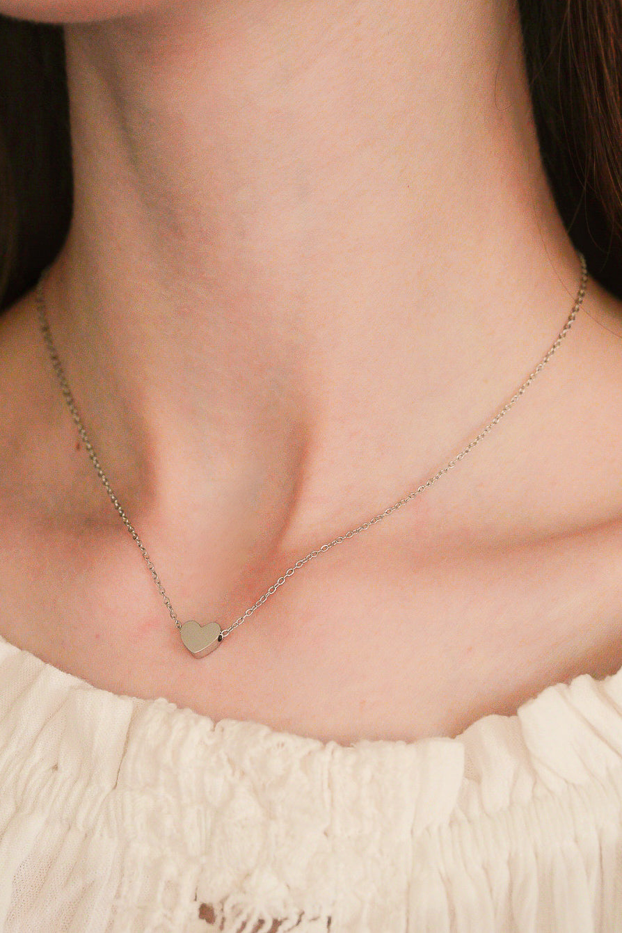 Sievietes kakla tuvplāns, uz kura valkā Como Jewelry Kaklarota - Sirds, smalku nerūsējošā tērauda kaklarotu ar sirsniņas piekariņu, kas veidots ar baltas krāsas, teksturētu, elastīgu kakla izgriezumu.