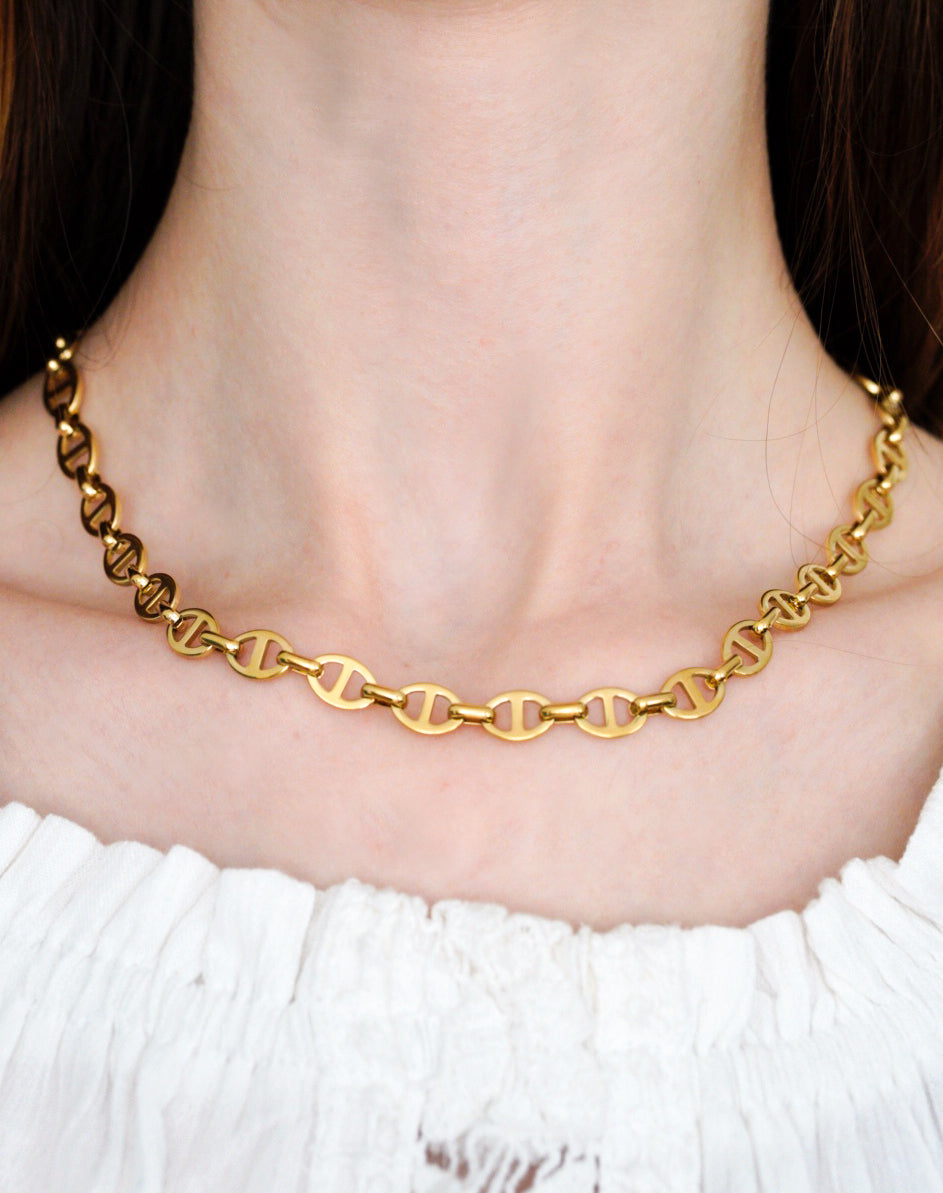 Persona, kas valkā Como Jewelry Kaklarota - Zelta ķēde ar ovālām, zirga koduma formas saitēm, kas kombinēta ar baltu, no pleciem novilktu fakturētu kreklu.