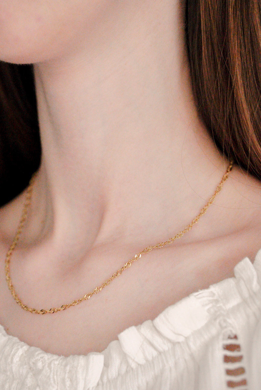 Sievietes kakla tuvplāns, uz kura redzama Como Jewelry Kaklarota - Zelta vijums, moderna zelta kaklarota, kas kombinēta ar baltu rūtainu kreklu. Viņas garie brūnie mati ierāmē maigi izplūdušo fonu.