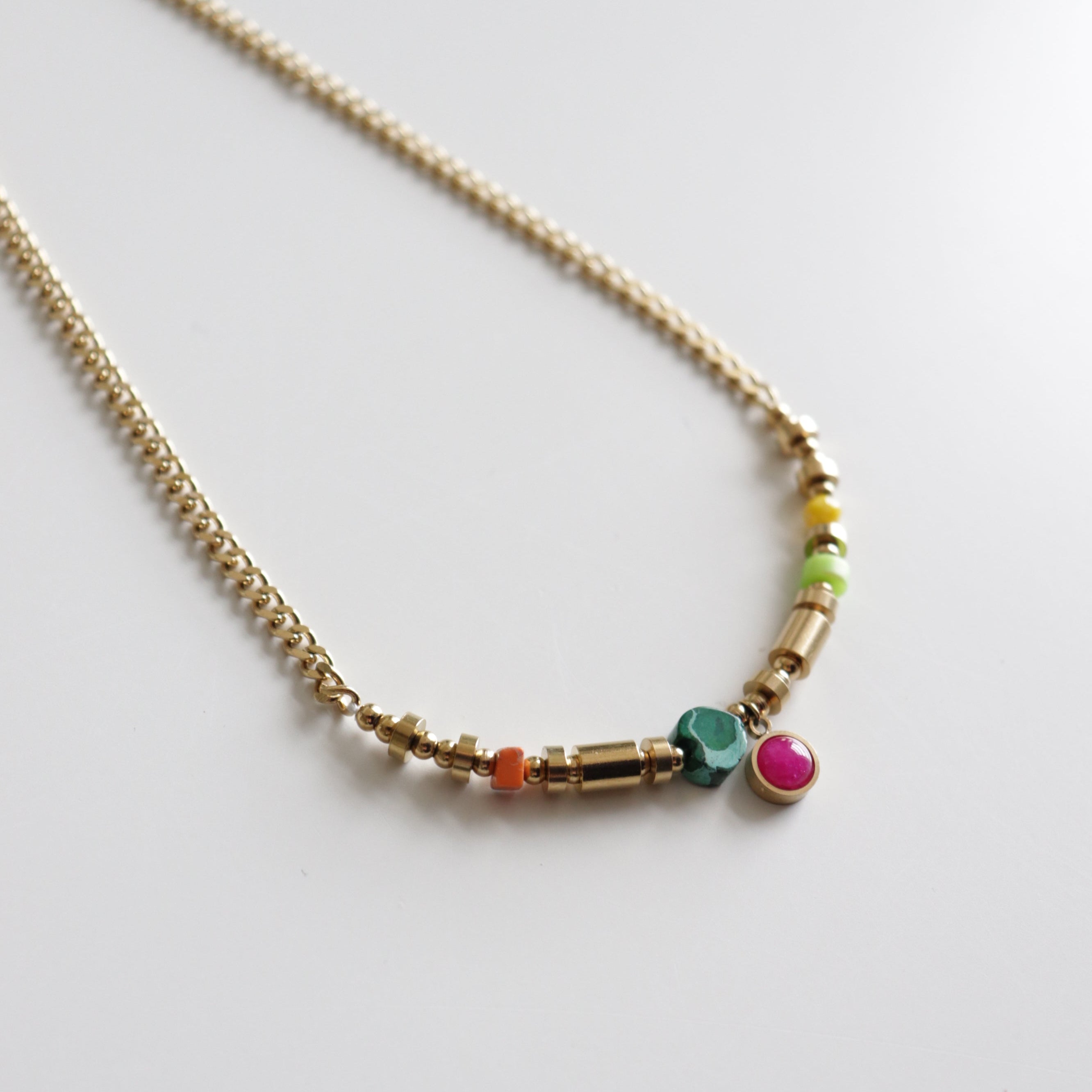 Kaklarota - Rio no Como Jewelry: moderna juveliertērauda kaklarota ar zelta ķēdi un asimetriski izvietotām daudzkrāsainām pērlītēm (zaļa, rozā, oranža, dzeltena un zelta), uz balta pamata.