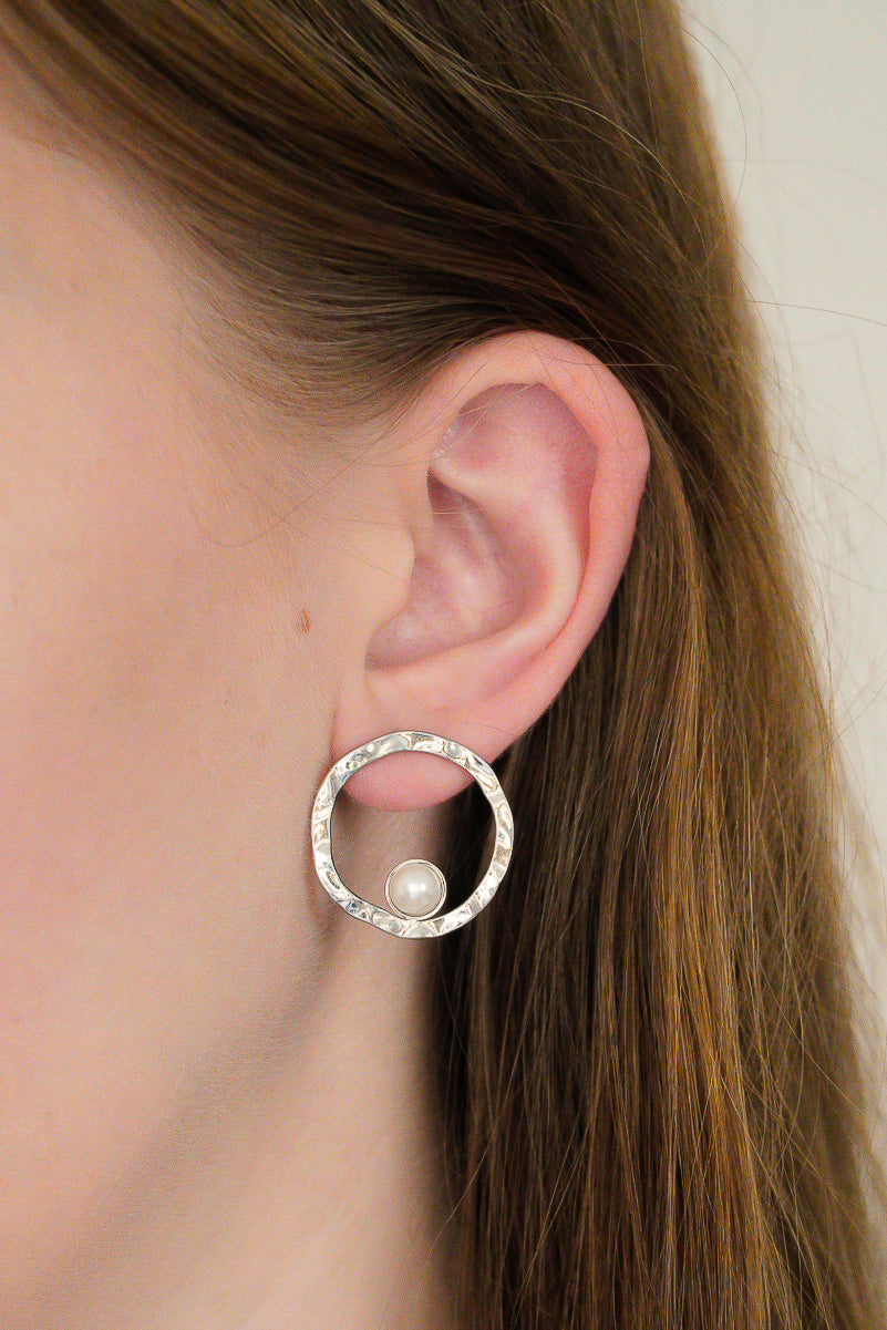 Detalizēts sievietes auss, kurai ir Como Jewelry Mazie auskari - Rietošā pērle: sudraba aproces ar faktūru un nelielu baltu pērli; viņas gaiši brūnie mati ir aizbīdīti aiz auss.