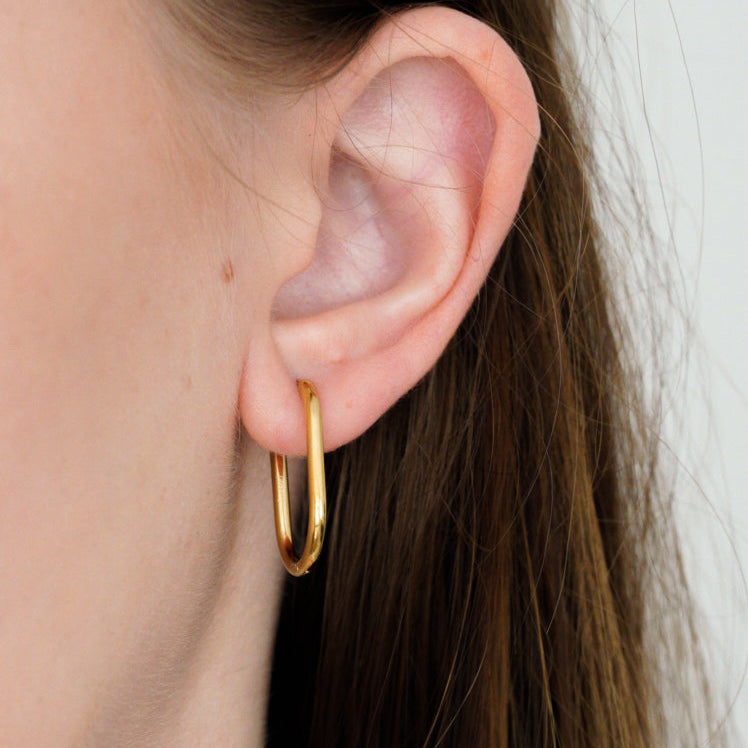 Tuvplāns cilvēka ausij ar taisniem brūniem matiem, kam ir Como Jewelry Mazie auskari - Ovāls. Vienkāršajos ovālajos auskaros ir zelta pārklājums. Fons ir vienkrāsains un gaišs.