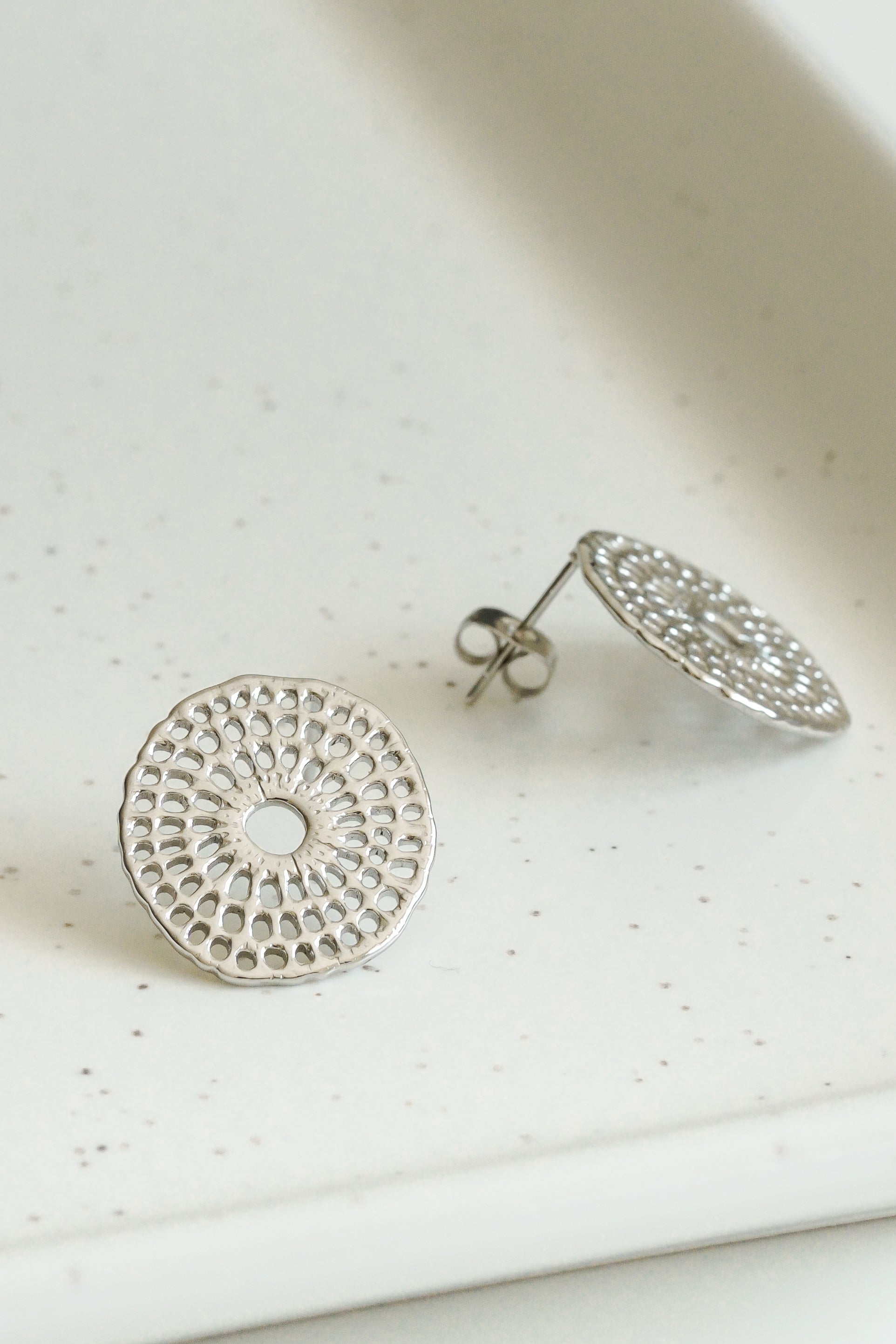 Como Jewelry Mazie auskari - Saules pinumi ar sarežģītiem apaļiem sudraba rakstiem, ko iedvesmojuši rokdarbu raksti, ir izvietoti uz baltas, raibas virsmas - viens auskariņš ir plakans, otrs - vertikāls, uz kura redzams stienis.