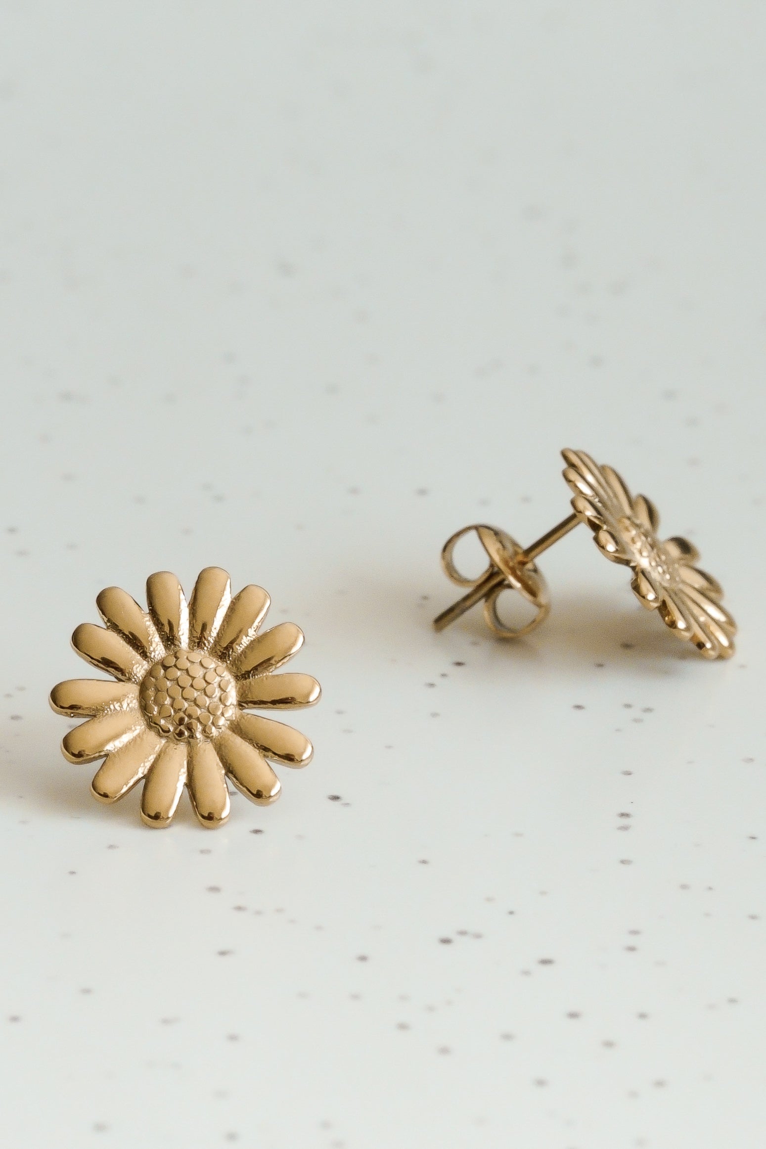 Como Jewelry Mazie auskari - Daisy: zelta pārklāta tērauda auskari ar teksturētiem centriem un gludām lapiņām, izlikti uz balta virsmas ar pelēkiem punktiņiem. Hipoalerģiski, eleganti ikdienas valkāšanai.