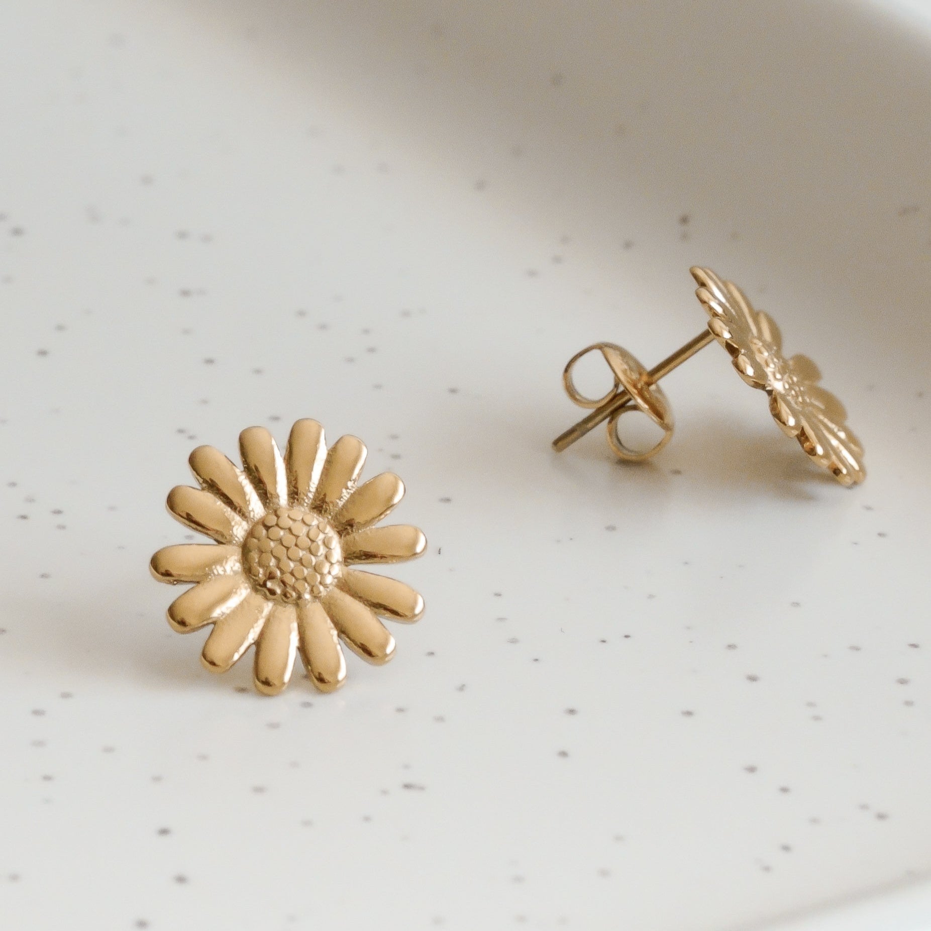 Mazie auskari pāris - Daisy by Como Jewelry, kas izgatavoti no zelta pārklāts tērauds, balstās uz baltas raibas keramikas virsmas, piedāvājot burvīgu un hipoalerģiski aksesuāru izvēli.