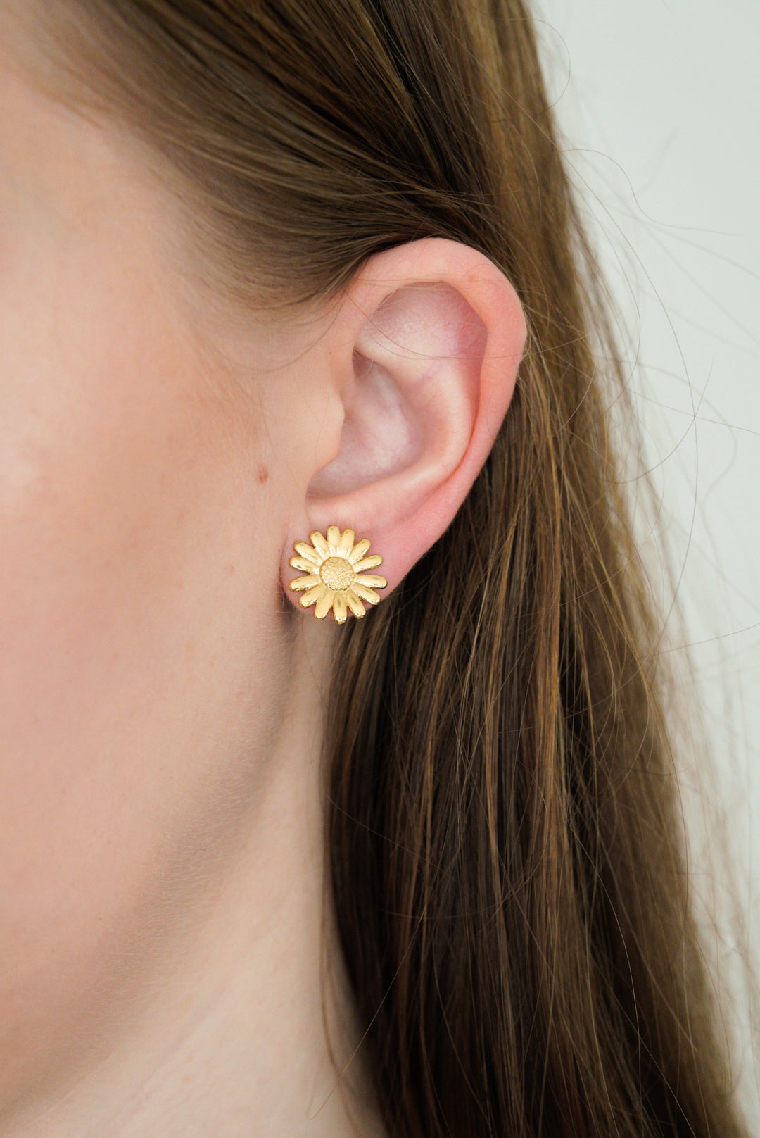 Tuvplāns personai ar taisniem, gaiši brūniem matiem, kurai ir Como Jewelry "Mazie auskari - Daisy" auskariņi, kas izgatavoti no hipoalerģiski zelta pārklāts tērauds. Gaiša āda un vienkrāsains, gaišs fons.