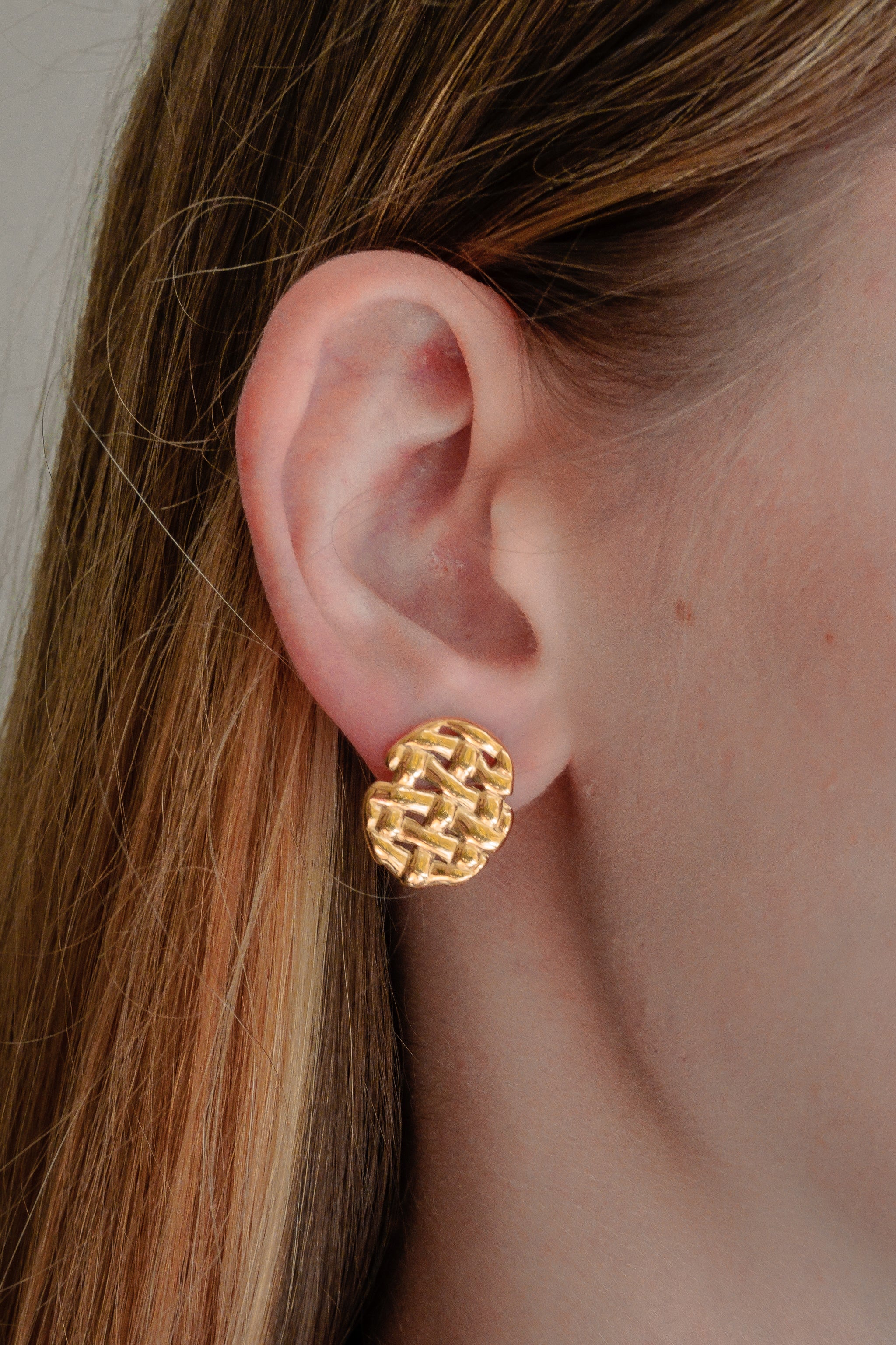 Tuvplānā redzama gaišādaina persona, kas valkā Mazie auskari - Pinums by Como Jewelry, ar apaļiem auskariem ar zelta austu tekstūru. Taisni, gaiši brūni mati ir glīti aizbīdīti aiz auss.