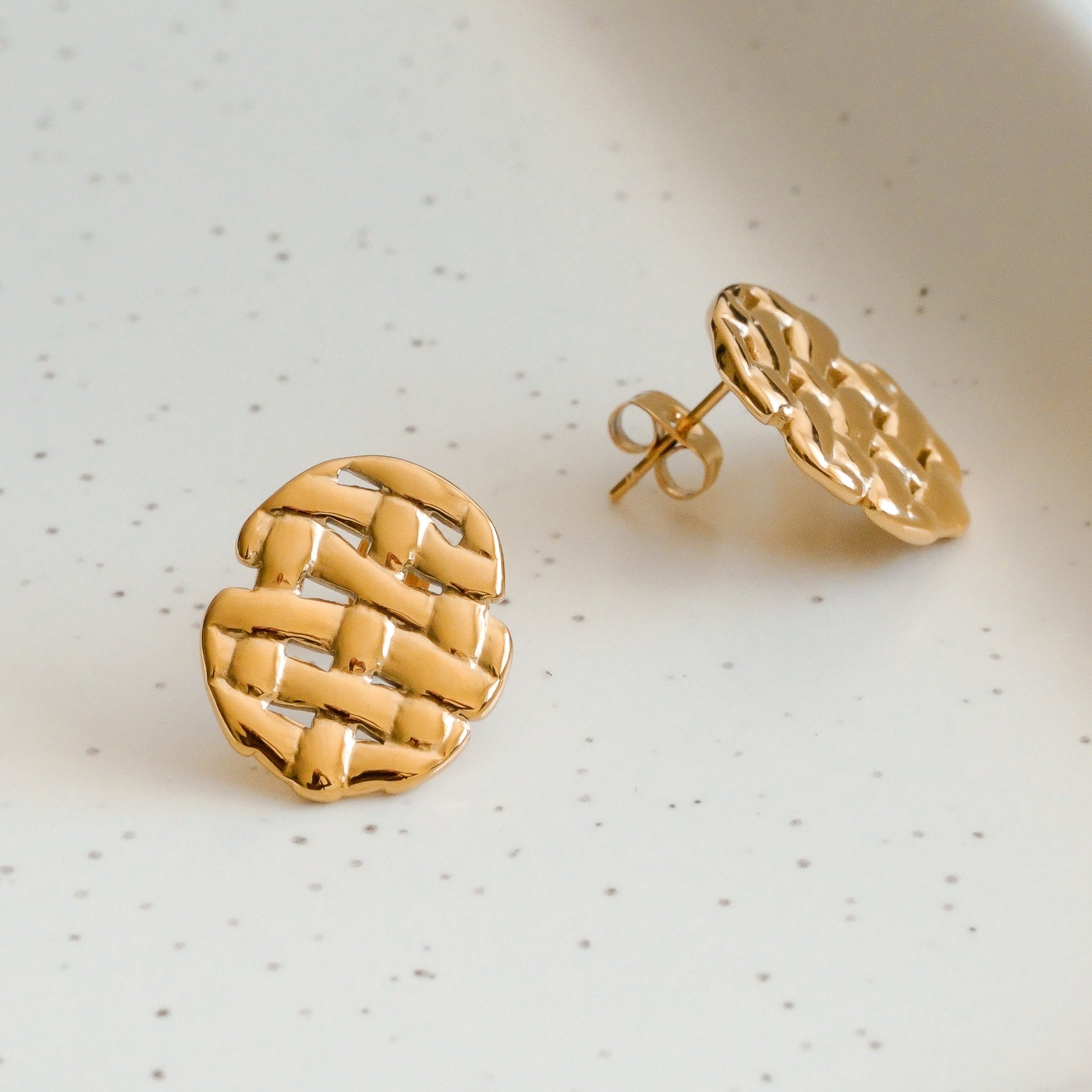 Mazie auskari - Pinums no Como Jewelry, ar zeltītu austu tekstūru un ovālu formu, balstās uz baltas keramikas virsmas ar plankumiem.