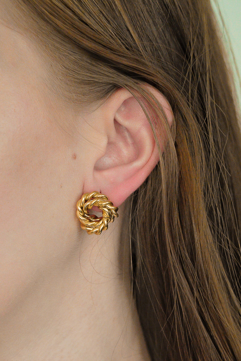 Sievietes auss tuvplānā, ko rotā Como Jewelry Mazie auskari - Mezgls, eleganti mezgla formas auskari. Viņas brūnie mati ir aizķērušies aiz auss, izceļot šos simboliskos elementus uz maiga, gaiša fona.