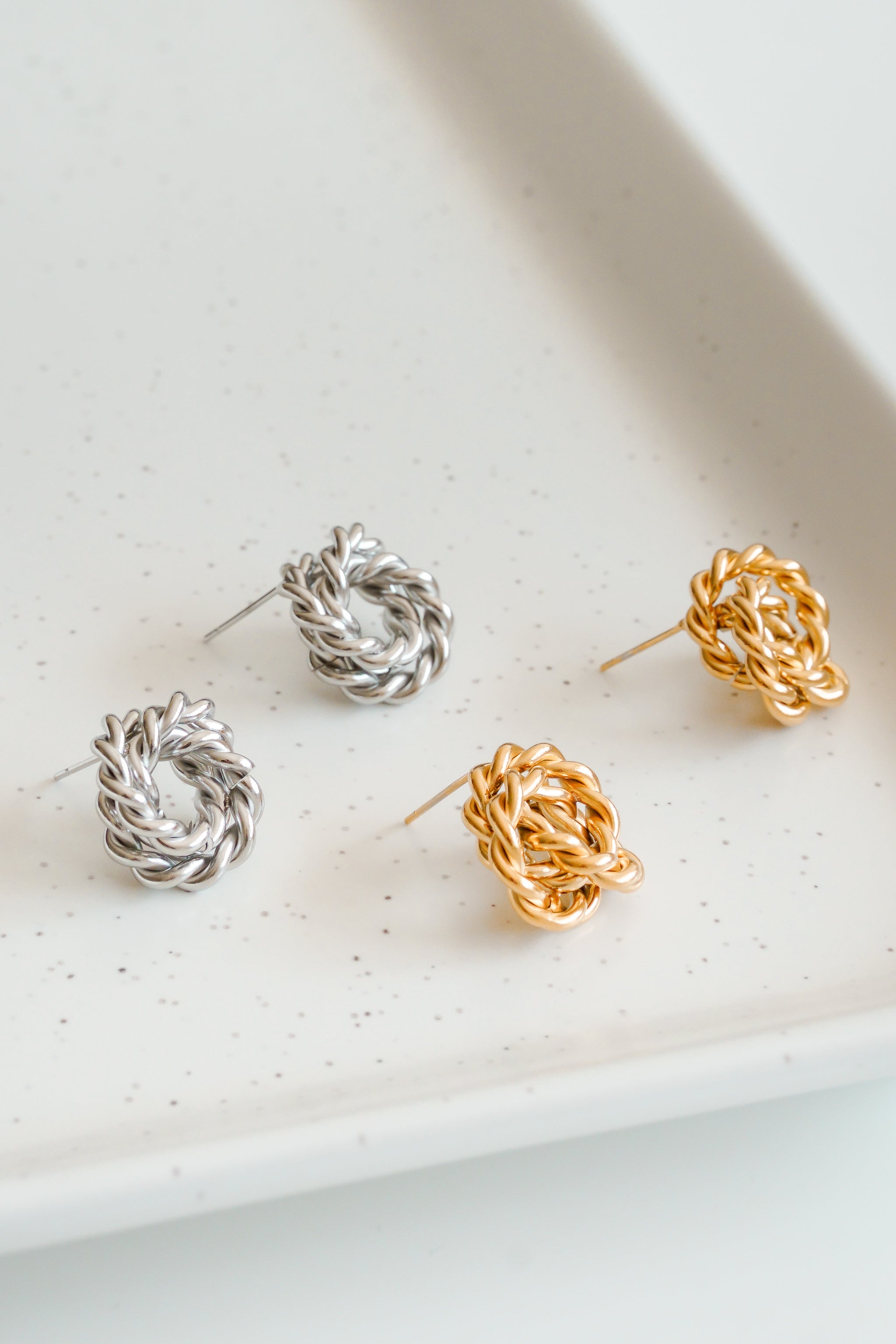 Divi pāri Mazie auskari - Mezgls by Como Jewelry - viens sudraba, otrs zelta - ir novietoti uz balta keramikas paplātes ar plankumiem. Šiem simboliskajiem rotaslietas elementiem ir eleganta zelta pārklāta tērauda auskari, kas ir pārlaicīga izskata.