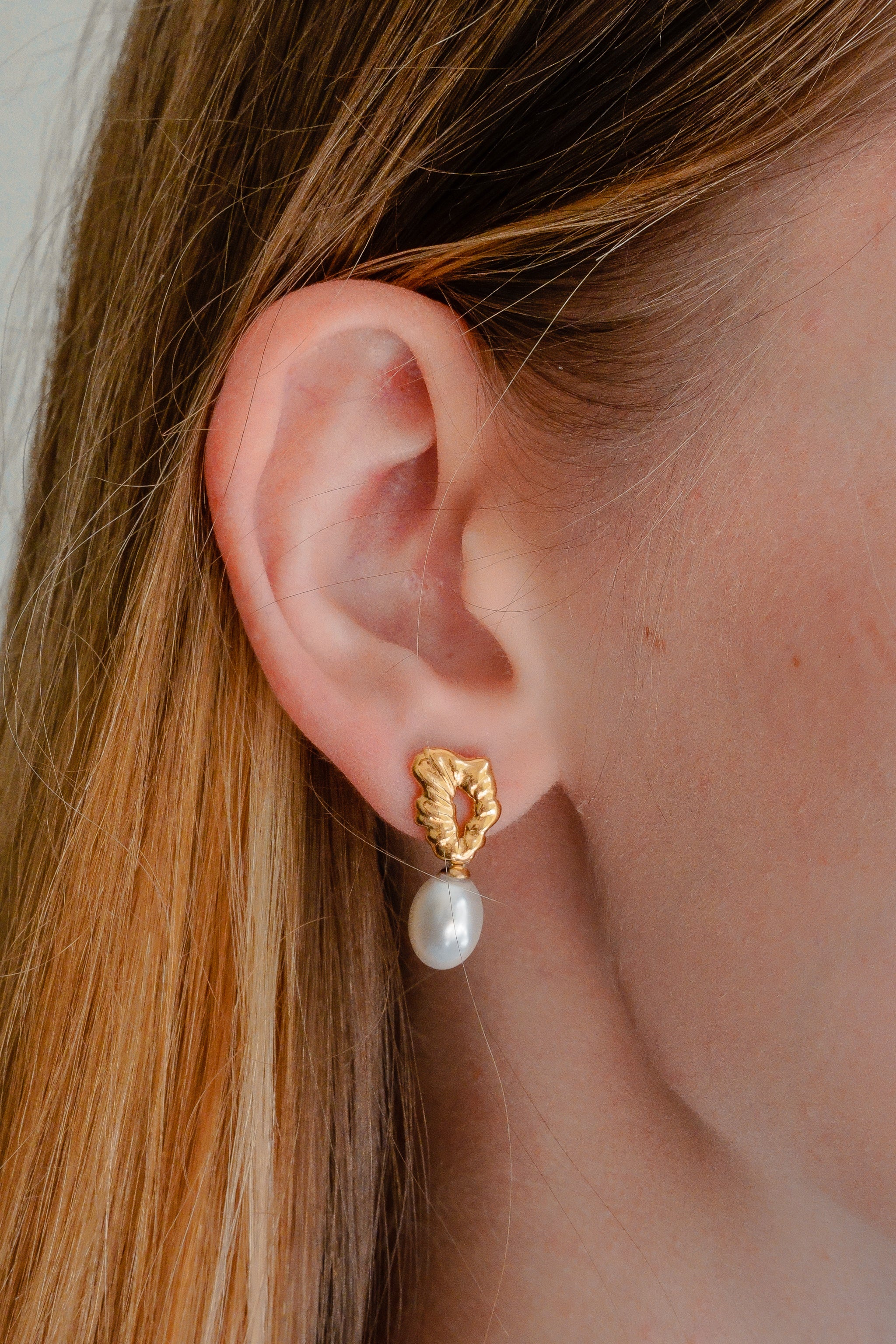 Detalizēts tuvplāns sievietes ausij ar Como Jewelry Mazie auskari - Plūstošā pērle ar teksturētu lapu dizainu un piekārtu baltu pērli. Izgatavoti no zelta pārklāta nerūsējošais tērauds, šie ūdensizturīgi auskari ir redzami ar matiem, kas ieķemmēti atpakaļ.