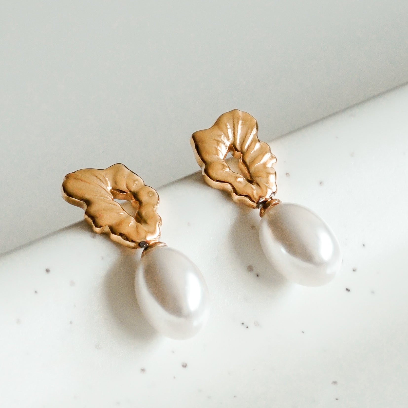 Como Jewelry Mazie auskari - Plūstošā pērle: zelta pārklāti nerūsējošā tērauda auskari ar teksturētiem, lapu formas akcentiem un ovālām baltām pērlēm, izstādīti uz balta, raibuma virsmas.