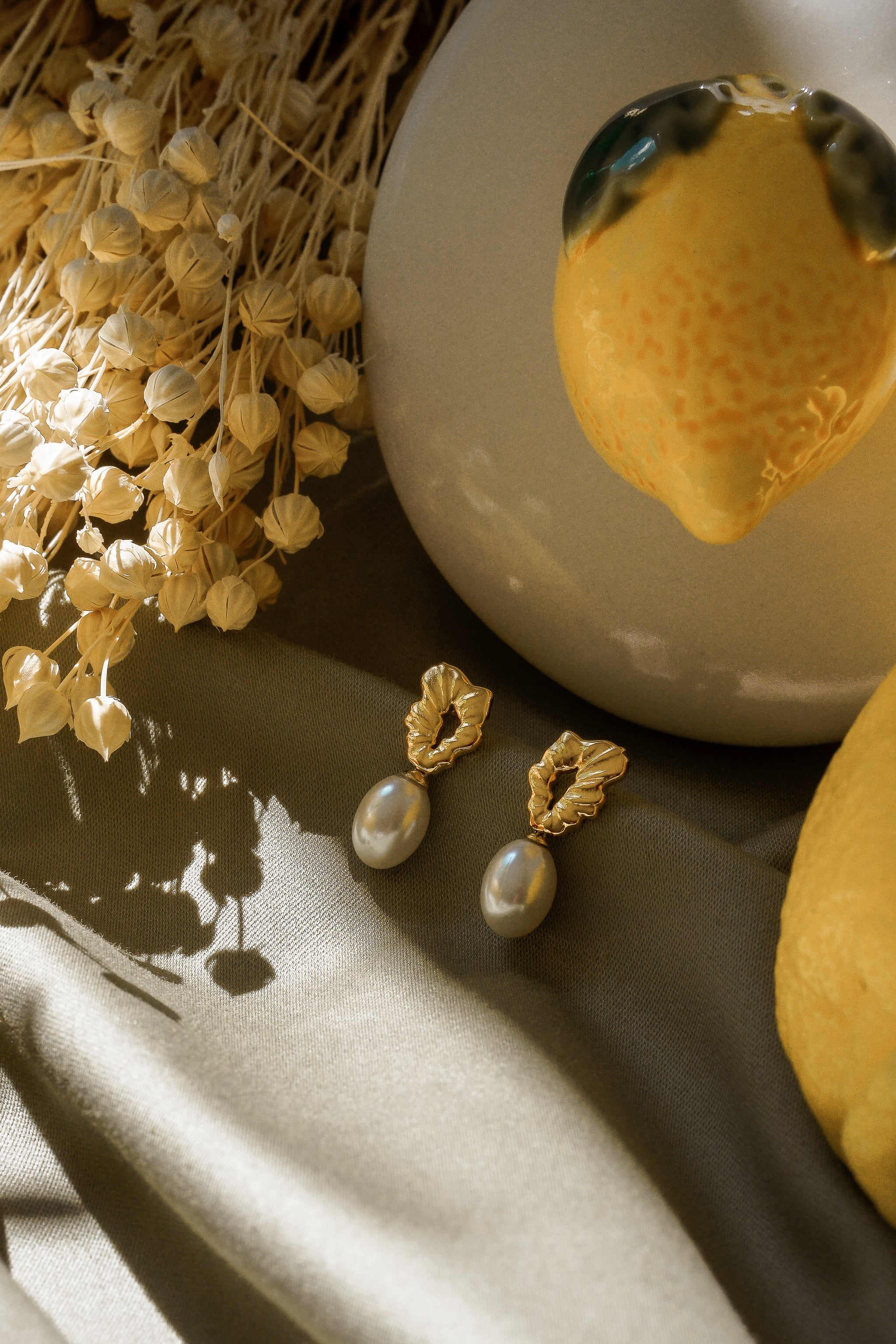 Mazie auskari - Como Jewelry plūstošā pērle ar apzeltītiem nerūsējošā tērauda lapu galiem, kas novietoti uz zaļgana auduma starp kaltētiem ziediem, keramikas citrona formas rotājumu un svaigu citronu.