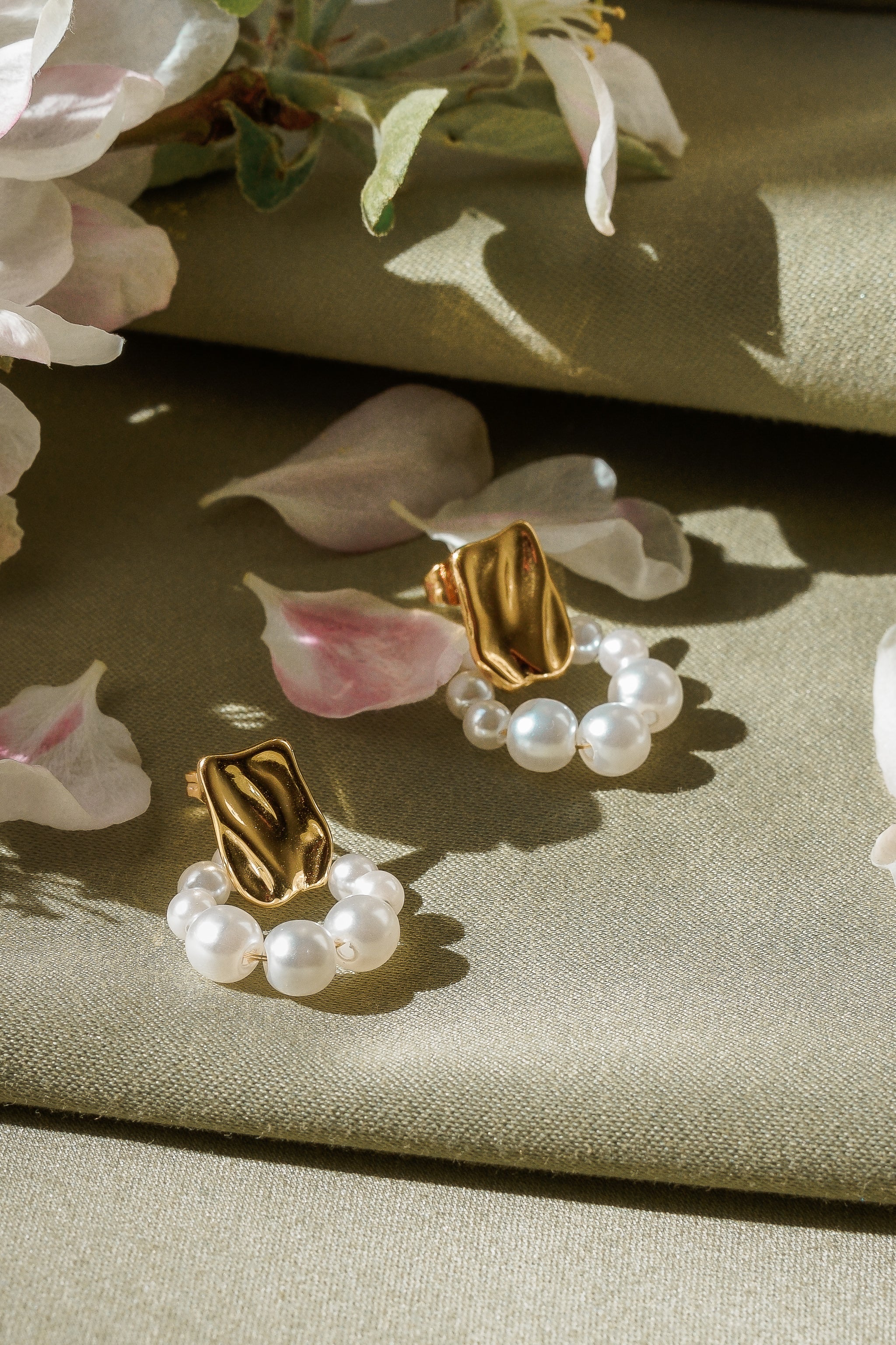 Como Jewelry Mazie auskari - Pērļu loki ar zeltītu pārklājumu un neregulārām formām, ko rotā mazas apaļas pērlītes, ir uzklāti uz gaiši olīvzaļa auduma starp maigi rozā ziedlapiņām un smalkiem ziediem.