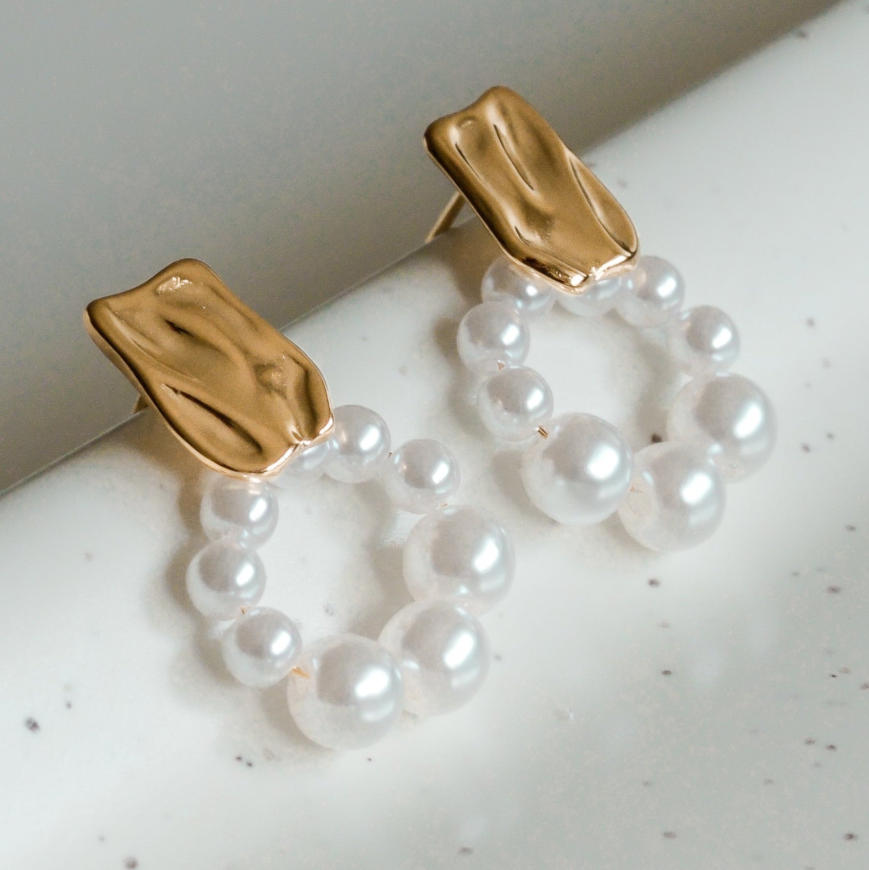 Mazie auskari - Pērļu loks by Como Jewelry: hipoalerģiski pērļu auskari ar neregulāras formas zelta pārklājuma aizdares un baltu pērļu lokiem, attēloti uz balta, raibuma virsmas. Ideāli jutīgām ausīm.