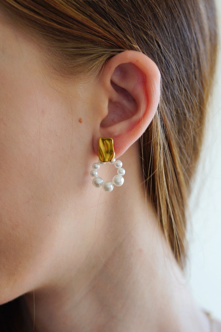 Detalizēts tuvplāns sievietes ausij ar Como Jewelry Mazie auskari - Pērļu loks: unikāli auskari ar apzeltītu taisnstūra stieni un apaļu baltu pērļu kārtojumu, attēlā ar brūniem matiem, kas novietoti aiz auss.