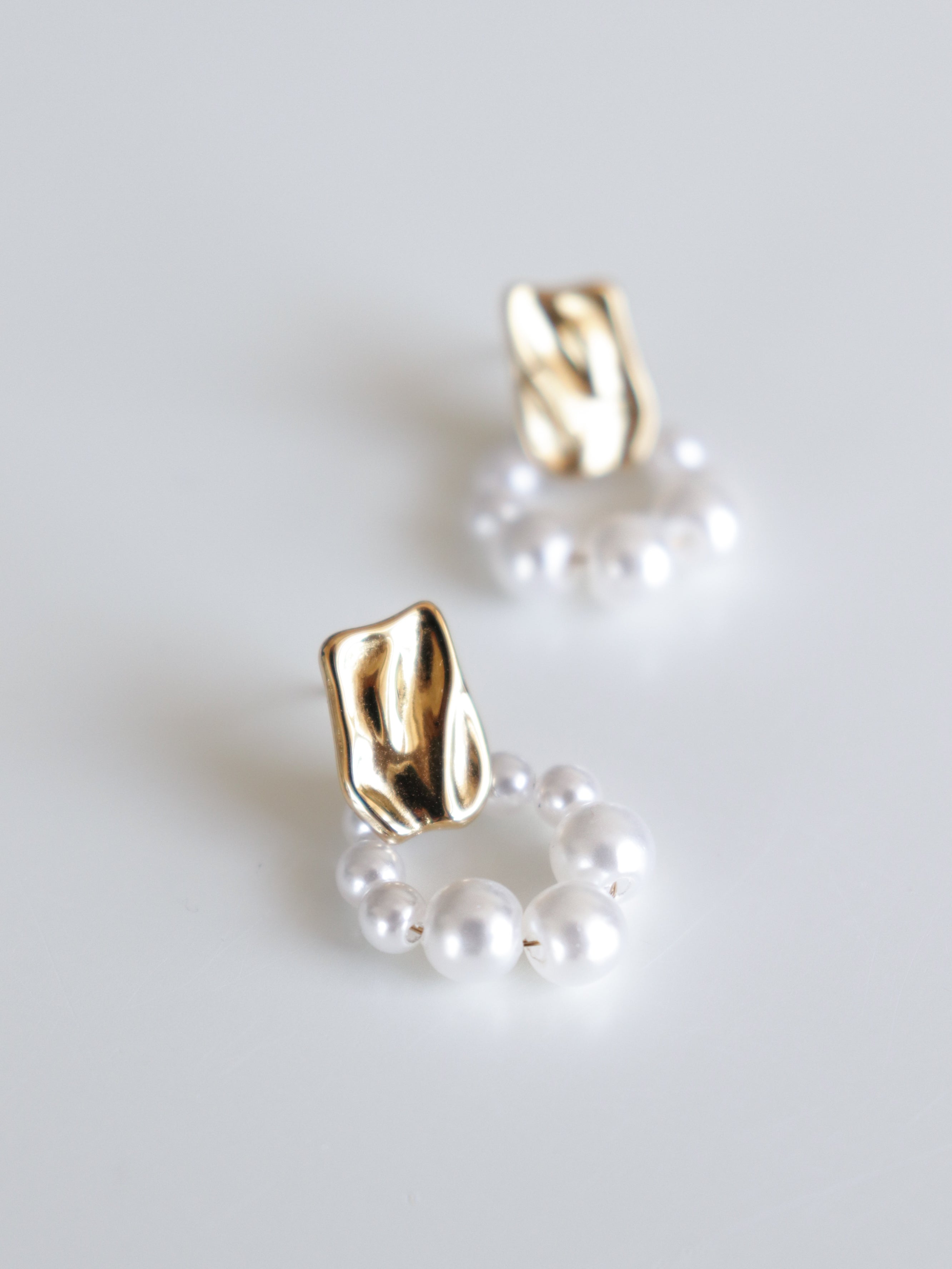 Como Jewelry Mazie auskari - Pērļu loks: neregulāras apzeltītas nagliņas ar apaļiem baltu pērļu dekoriem, kas izvietoti uz gludas, baltas virsmas.