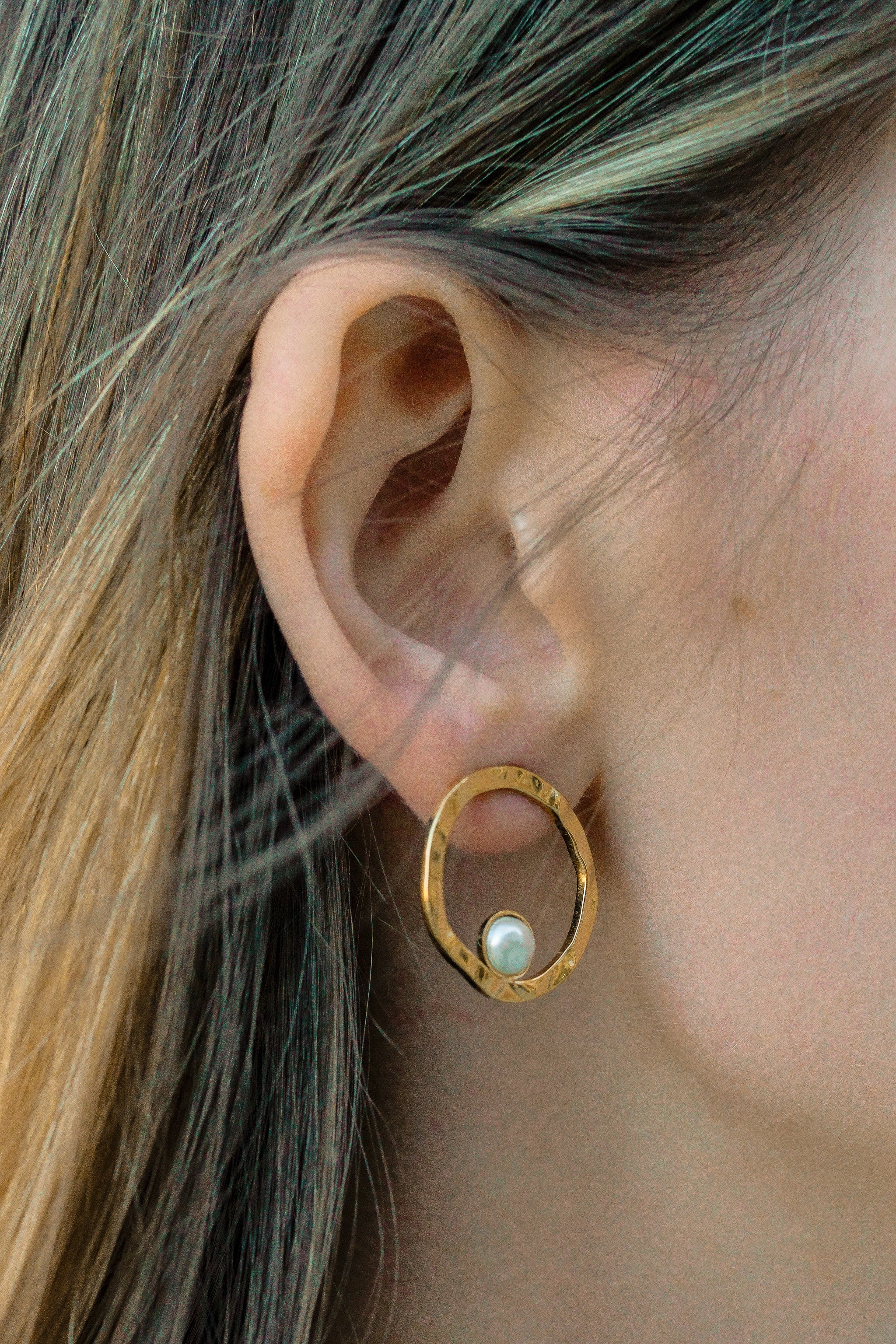 Tuvplāns sievietes ausij ar taisniem, gaiši brūniem matiem, kurai ir Mazie auskari - Rietošā pērle by Como Jewelry-moderni zelta riņķveida auskari ar mazu baltu pērli apļa iekšpusē.