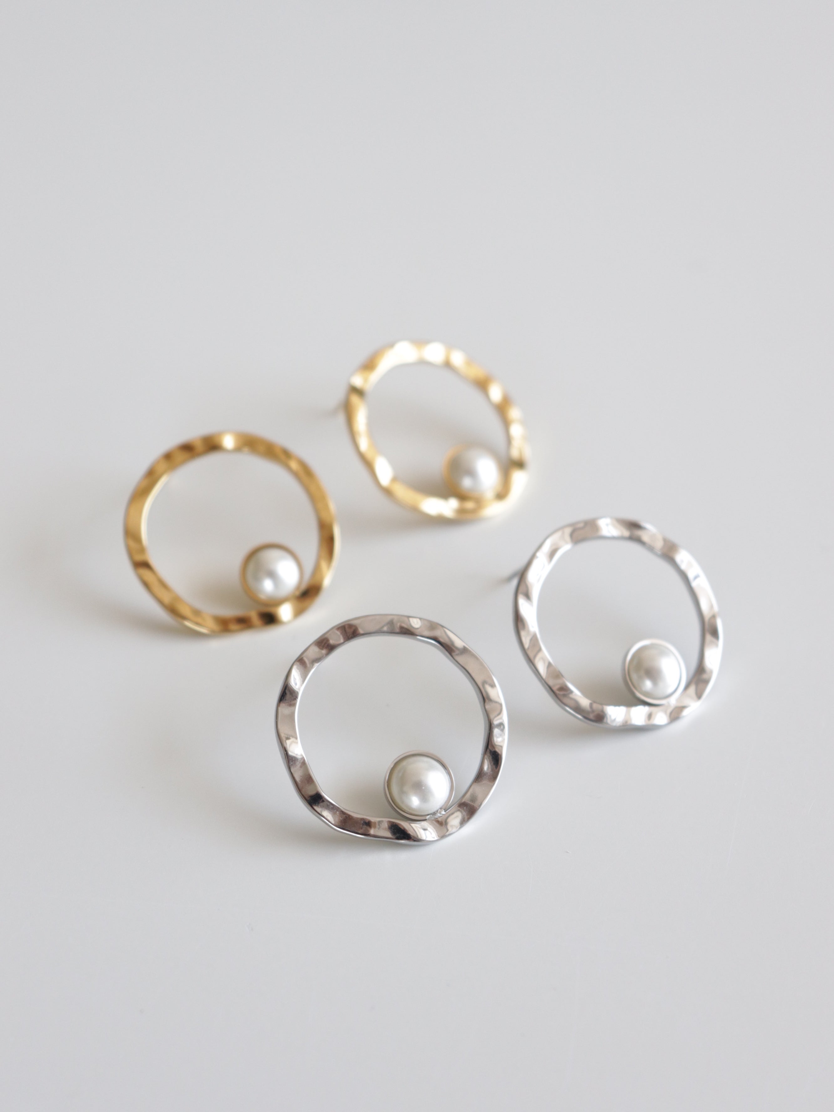 Mazie auskari "Rietošā pērle" no Como Jewelry-viens zelta, otrs sudraba tonī-ar āmurētu apli un pērli lejasdaļā eleganti izcelti uz balta fona.