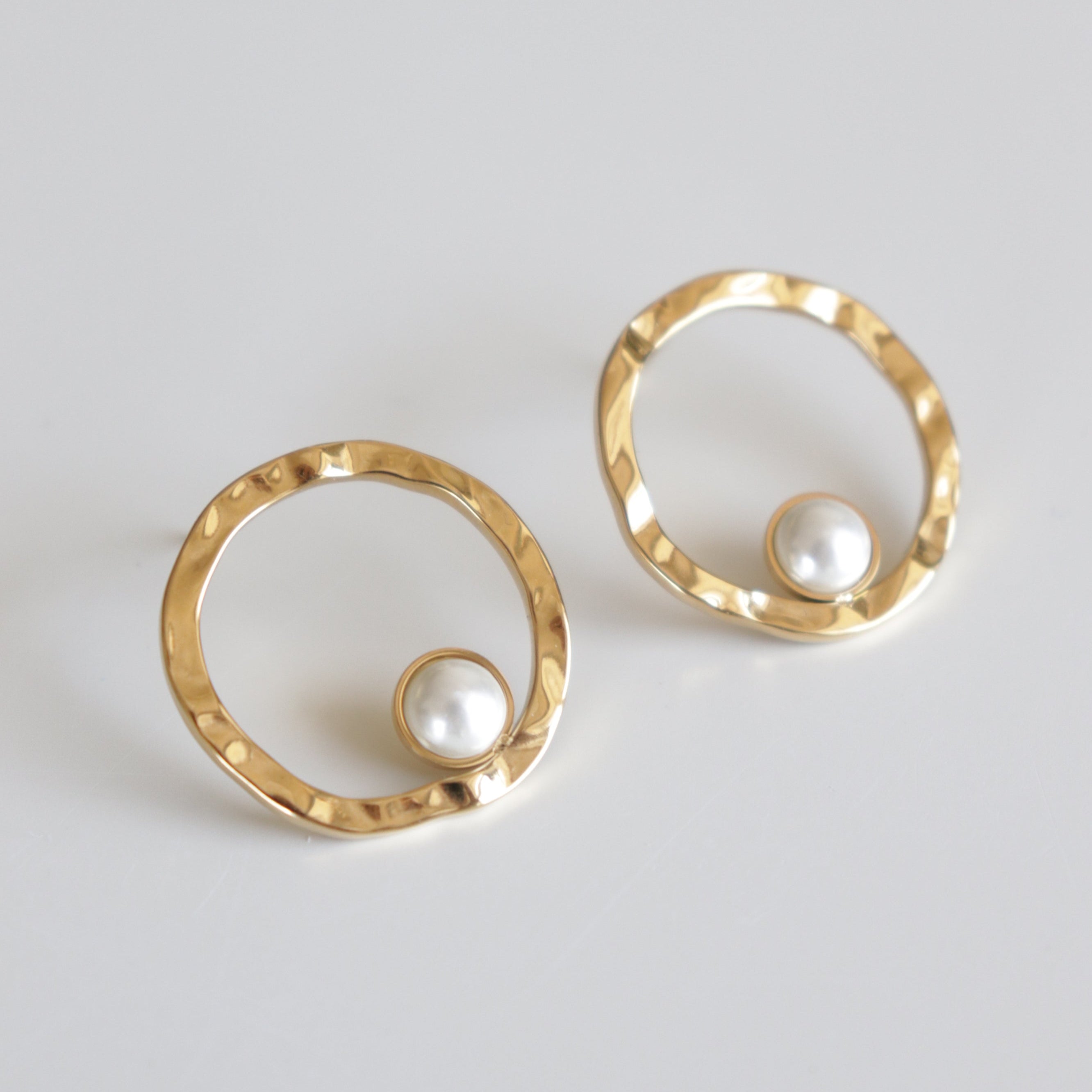 Mazie auskari - Rietošā pērle by Como Jewelry: zelta pārklājuma auskari ar kaltu tekstūru un baltu pērli apļa lejasdaļā, izstādīti uz balta fona.