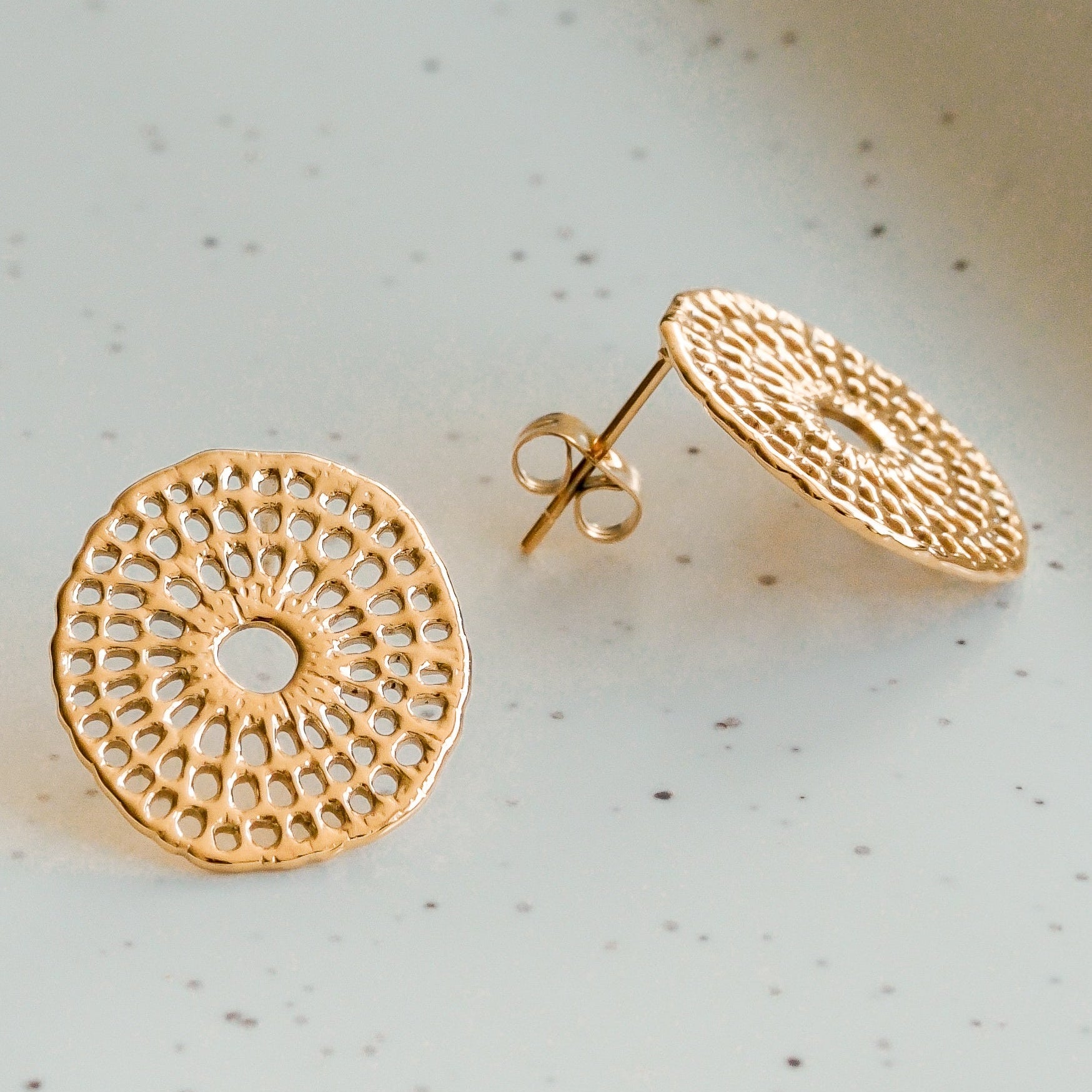 Divi apaļi, zeltīti, teksturēti kniedes auskari - "Mazie auskari - Saules pinums" - ar ažūriem mežģīņu mežģīņu rokdarbu raksti balstās uz gaišas raibas virsmas, no kuriem viens ir plakans, bet otrs - vertikāls, lai parādītu stieni.