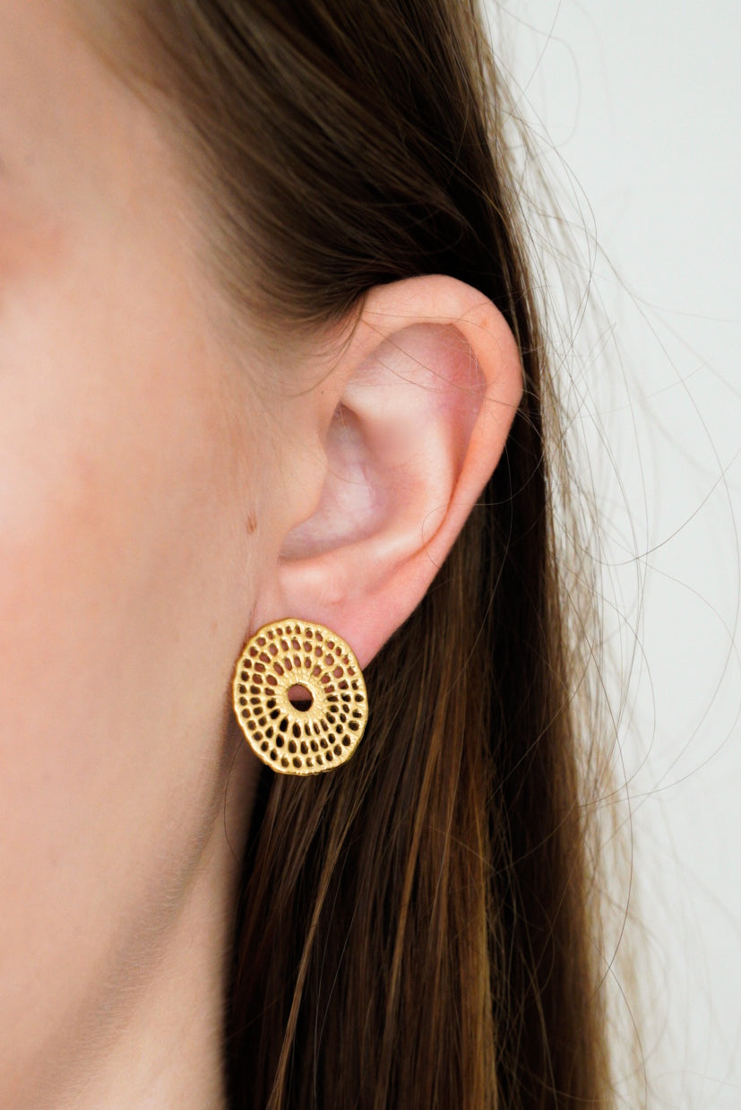 Tuvplāns uz personas auss, kurai ir Como Jewelry Mazie auskari - Saules pinums, ar nelielu, apaļu zelta auskaru ar izgrieztu apaļu rakstu un zelta pārklājumu. Gar sejas sānu daļēji redzami brūnie mati.