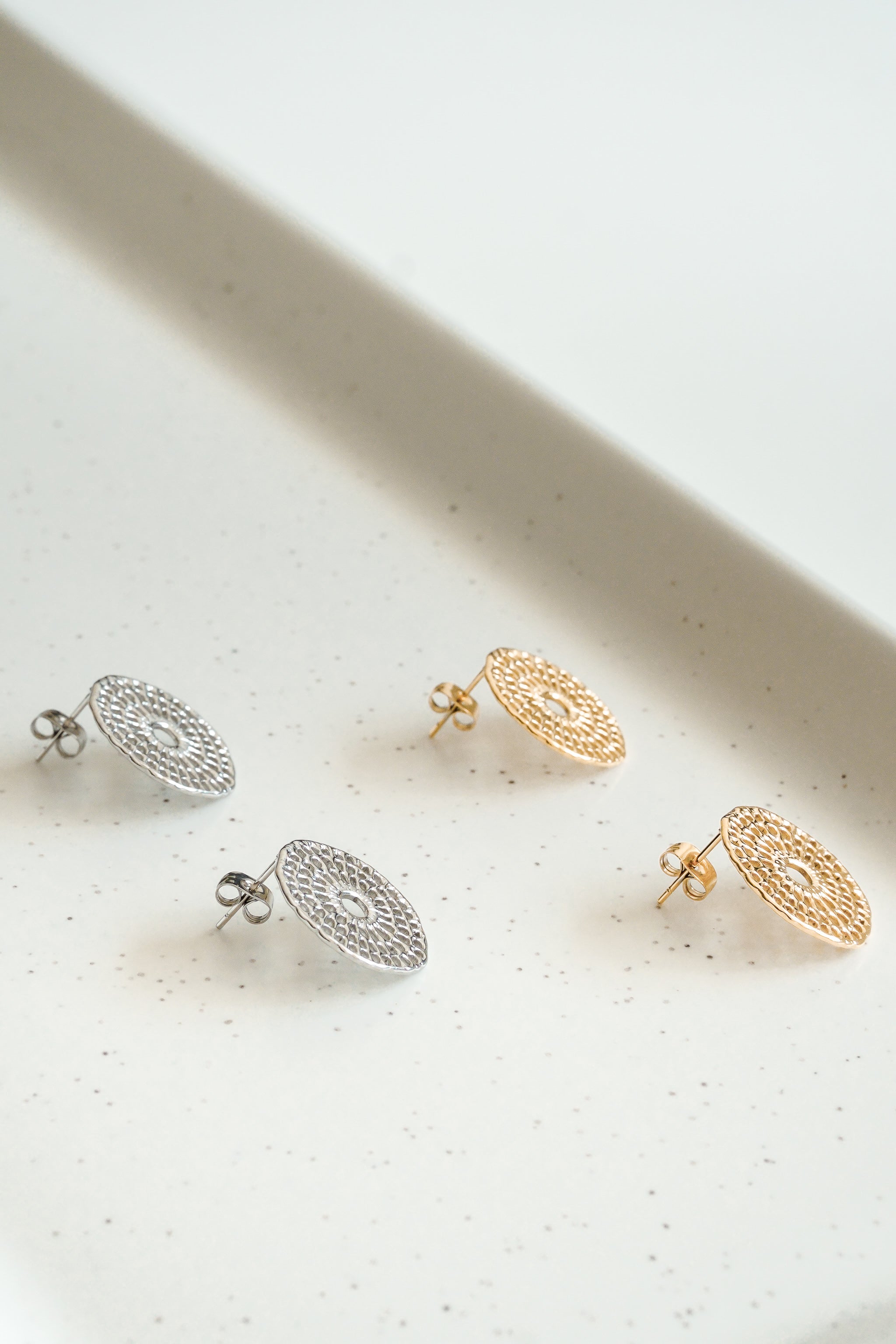 Divi pāri Mazie auskari - Saules pinumi no Como Jewelry, iedvesmojoties no rokdarbu raksti, ir izvietoti uz raibas baltas virsmas. Katrai tapai ir plakans, apaļš mežģīnēm līdzīgs sudraba vai zelta pārklājums.