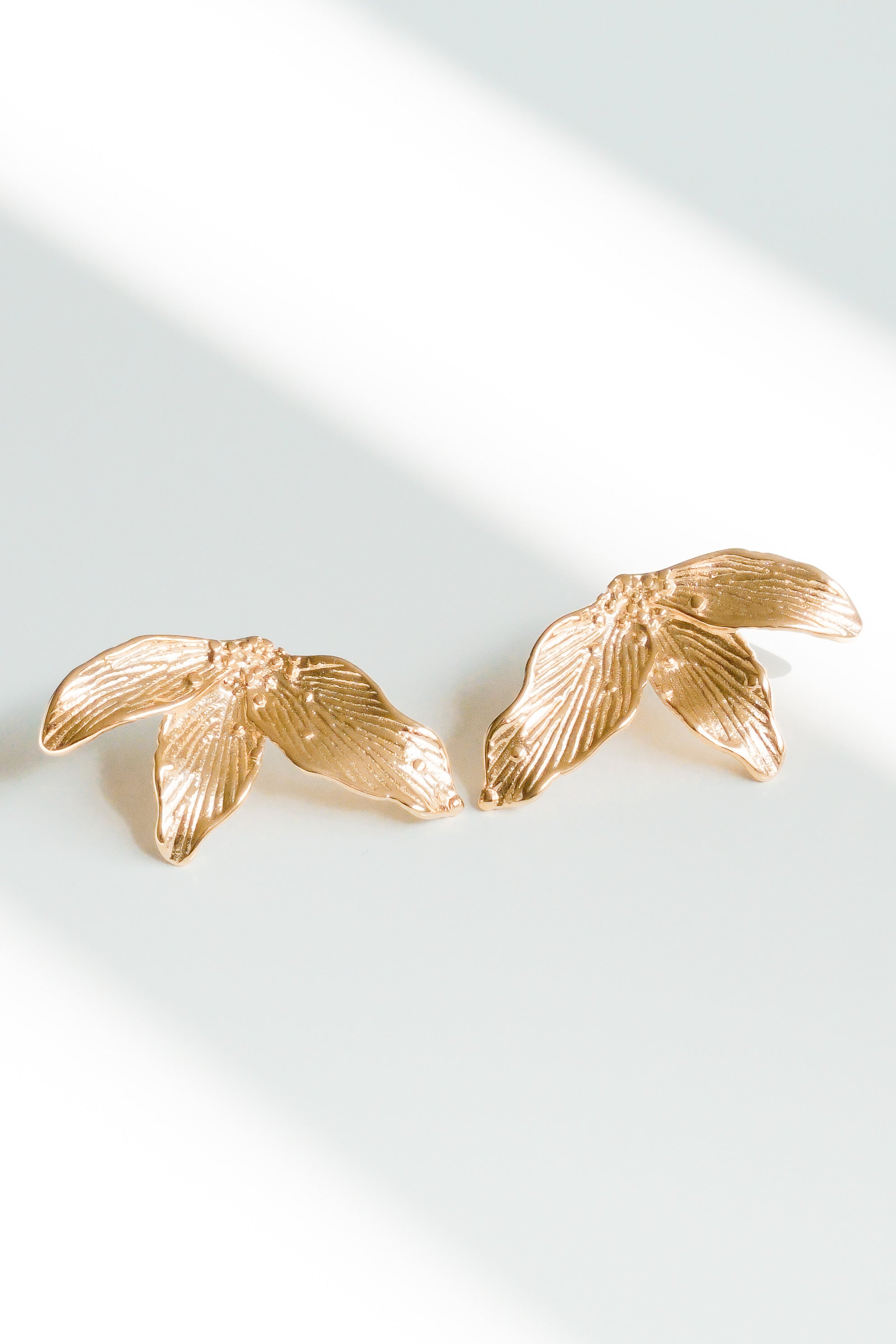 Mazie auskari - Ziedlapiņas no Como Jewelry, kas izgatavoti no zelta pārklāta nerūsējošais tērauds, balstās uz baltas virsmas ar lapu iedvesmotām formām. Šos elegantos hipoalerģiskos auskari izceļ diagonāli gaismas un ēnu raksti.