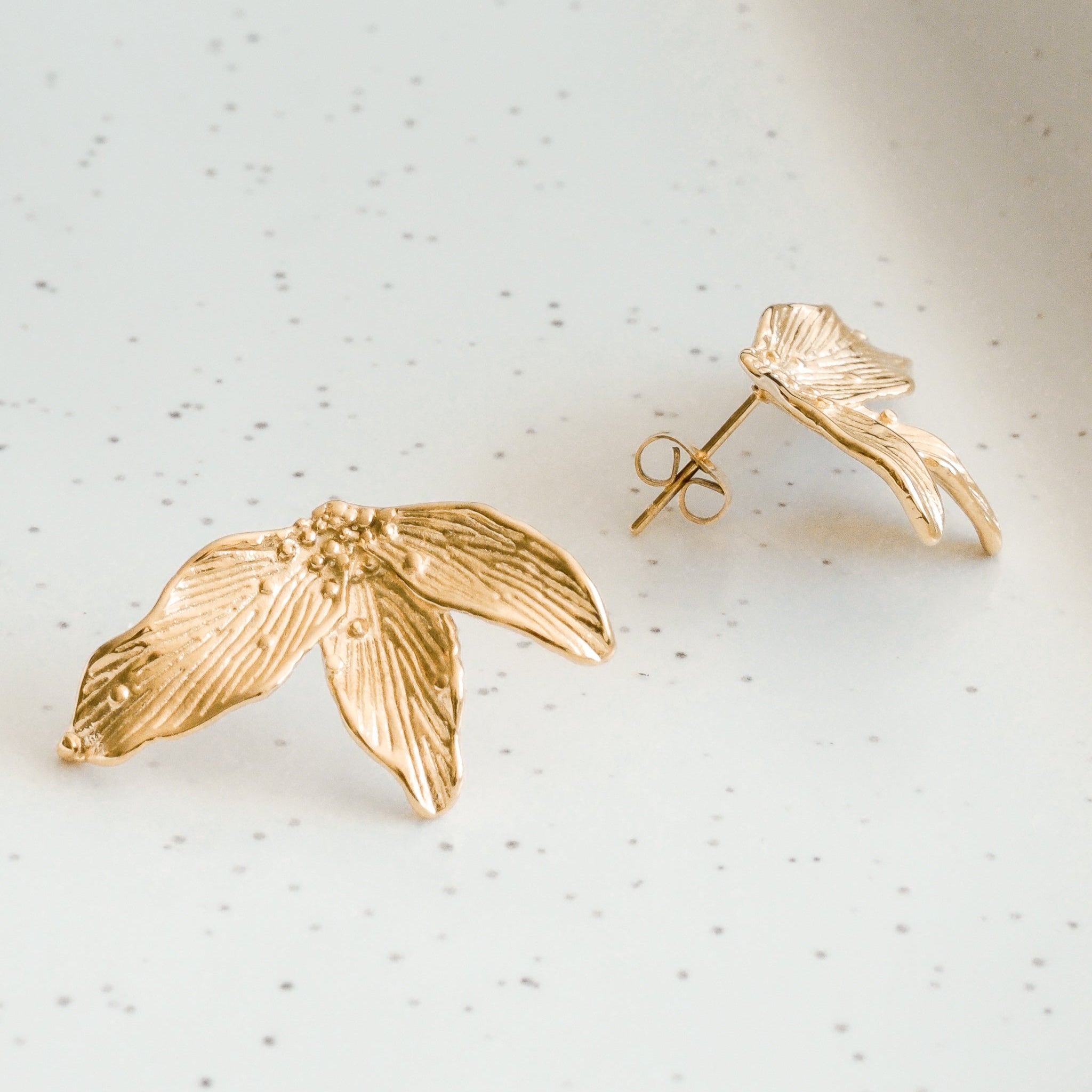 Mazie auskari - Como Jewelry Ziedlapiņas, kas izgatavotas no zelta pārklājuma nerūsējošais tērauds un veidotas kā maigas lapiņas, ir izvietotas uz baltas raibas virsmas - viena plakana, otra vertikāla, parādot tapas aizmuguri.