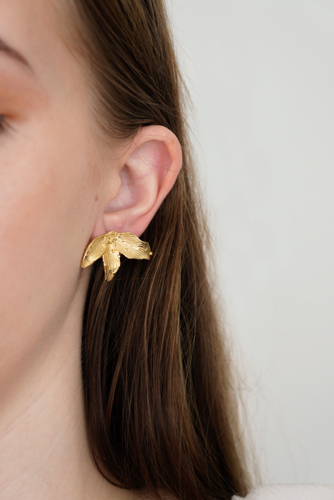 Sievietes auss tuvplānā ar gariem brūniem matiem, kurai ir Mazie auskari - Ziedlapiņas by Como Jewelry. Mazie auskari ir ziedlapiņu formā. Fons ir vienkrāsains un gaišs.