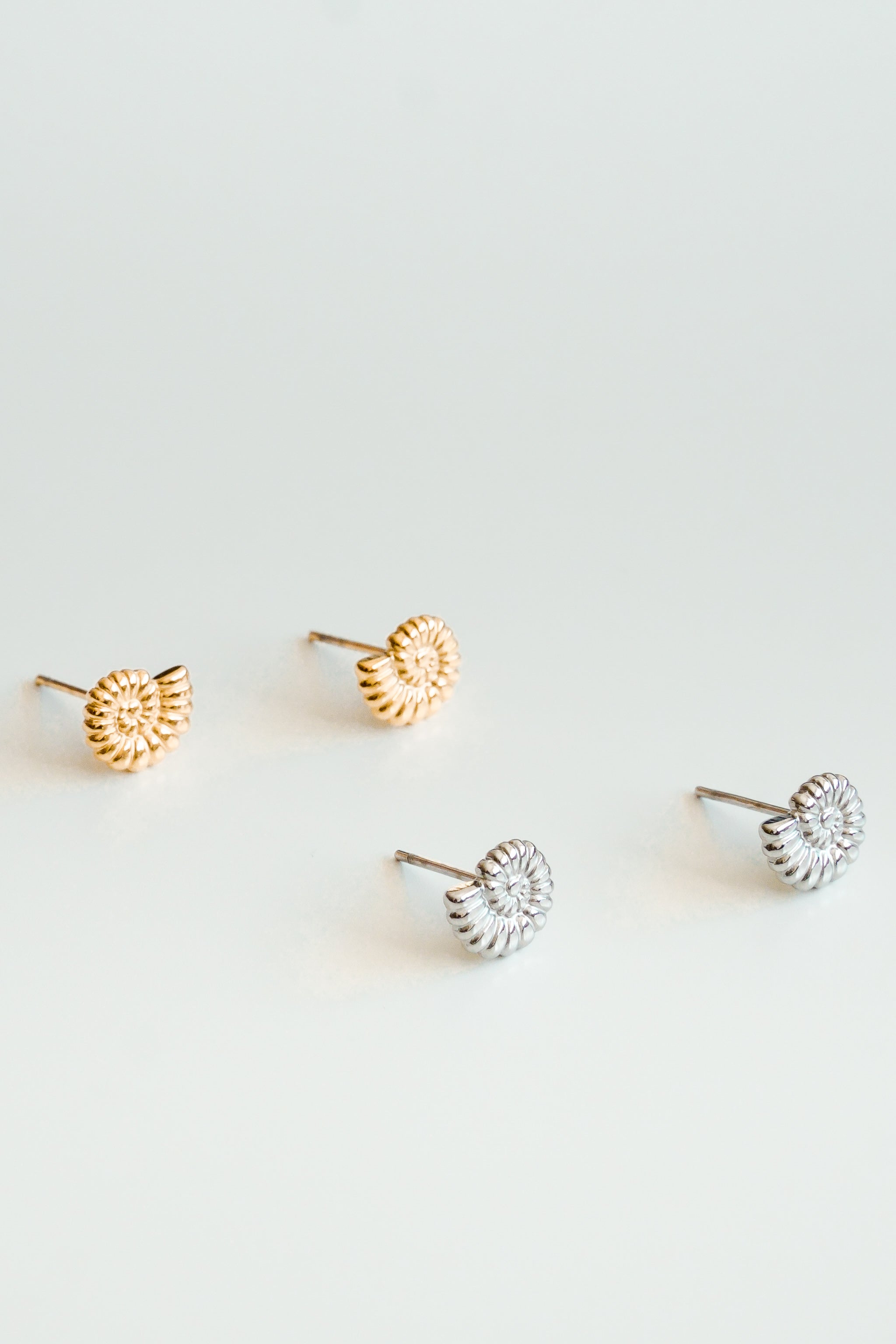 Izstādīti četri Como Jewelry "Mazie auskari - Gliemežvāki" - mazi, apaļi gliemežvāki ar izliektu ziedu rakstu: divi zelta pārklājuma (zelta tonī) un divi sudraba krāsā uz gaiša fona.