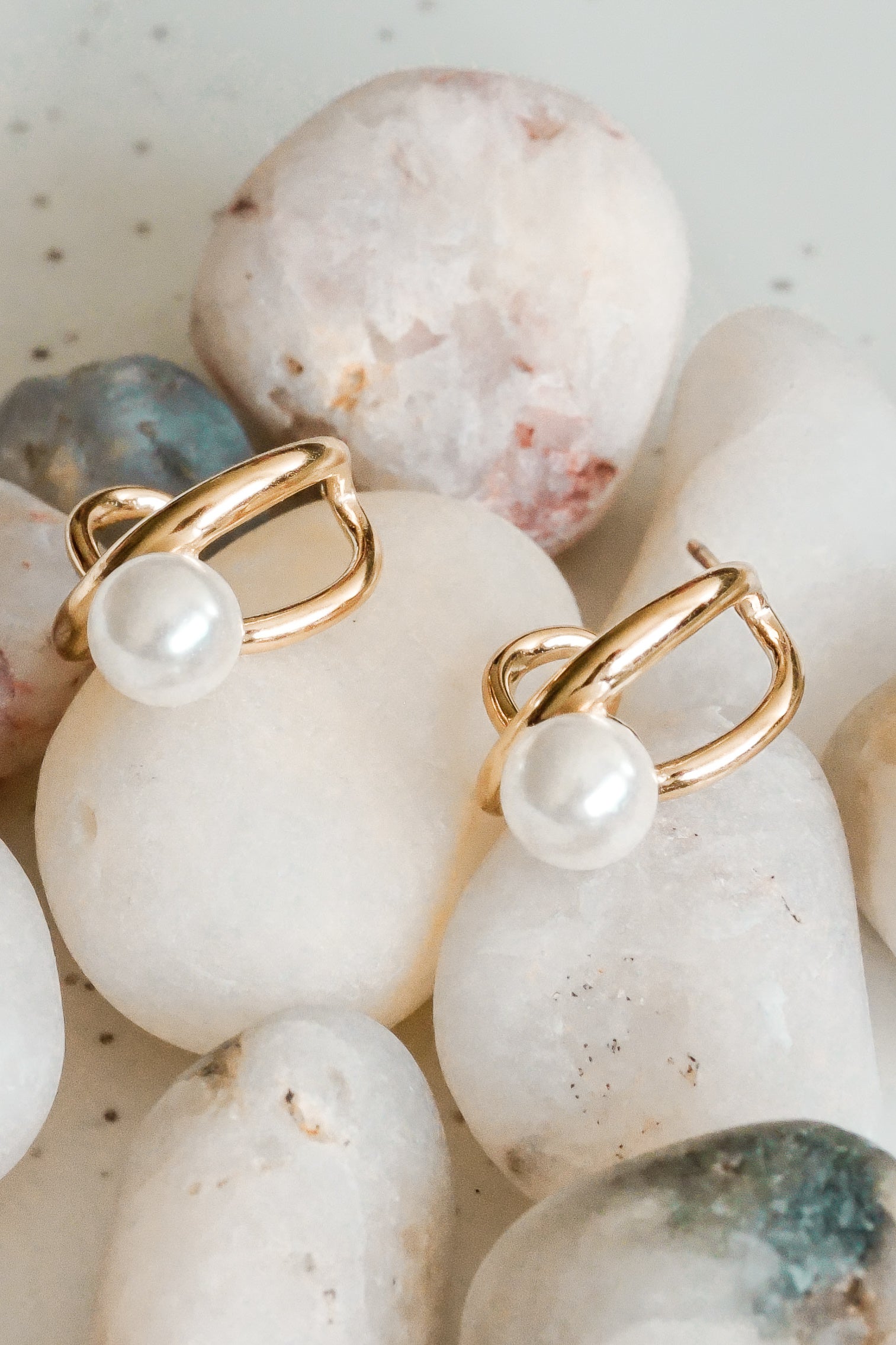 Mazie auskari - Loop no Como Jewelry ar 14k zelta pārklājumu un baltām imitācijas pērlēm eleganti atpūšas uz gludām, baltām oļiem.