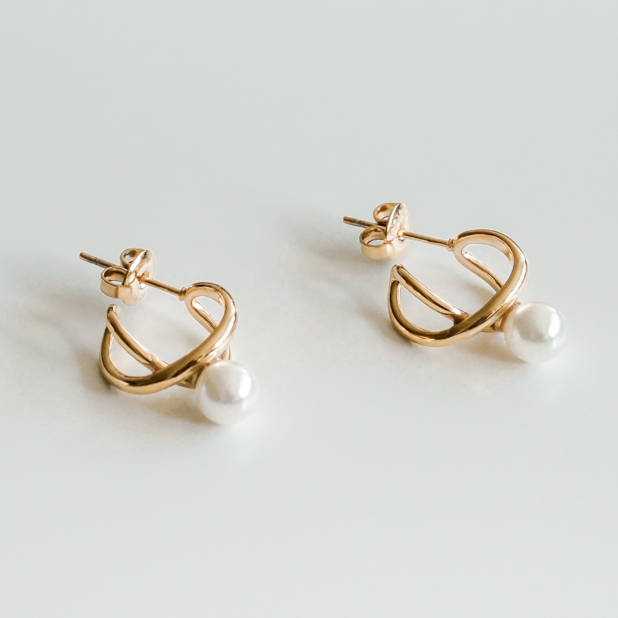 Mazie auskari - Loop by Como Jewelry ir 14k apzeltīti riņķveida auskari ar abstraktu savītu dizainu, katru no tiem rotā viena mākslīgā pērle pie pamatnes, kas attēlota uz balta fona.