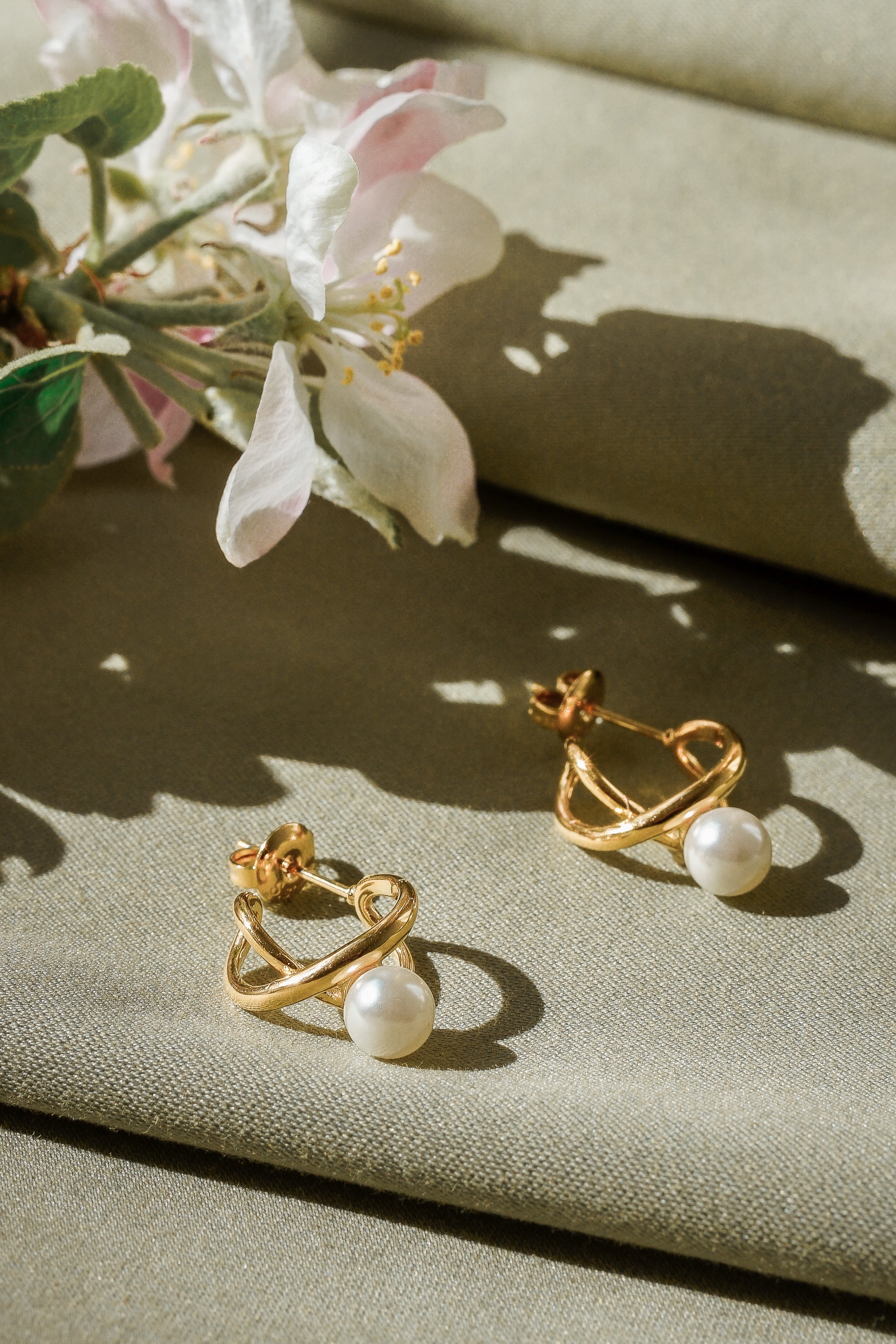 Mazie auskari - Loop no Como Jewelry ar 14k zelta pārklājumu un imitācijas pērļu kuloniem mirdz uz gaiši zaļa auduma, blakus maigām baltām un rozā puķēm dabīgā apgaismojumā.