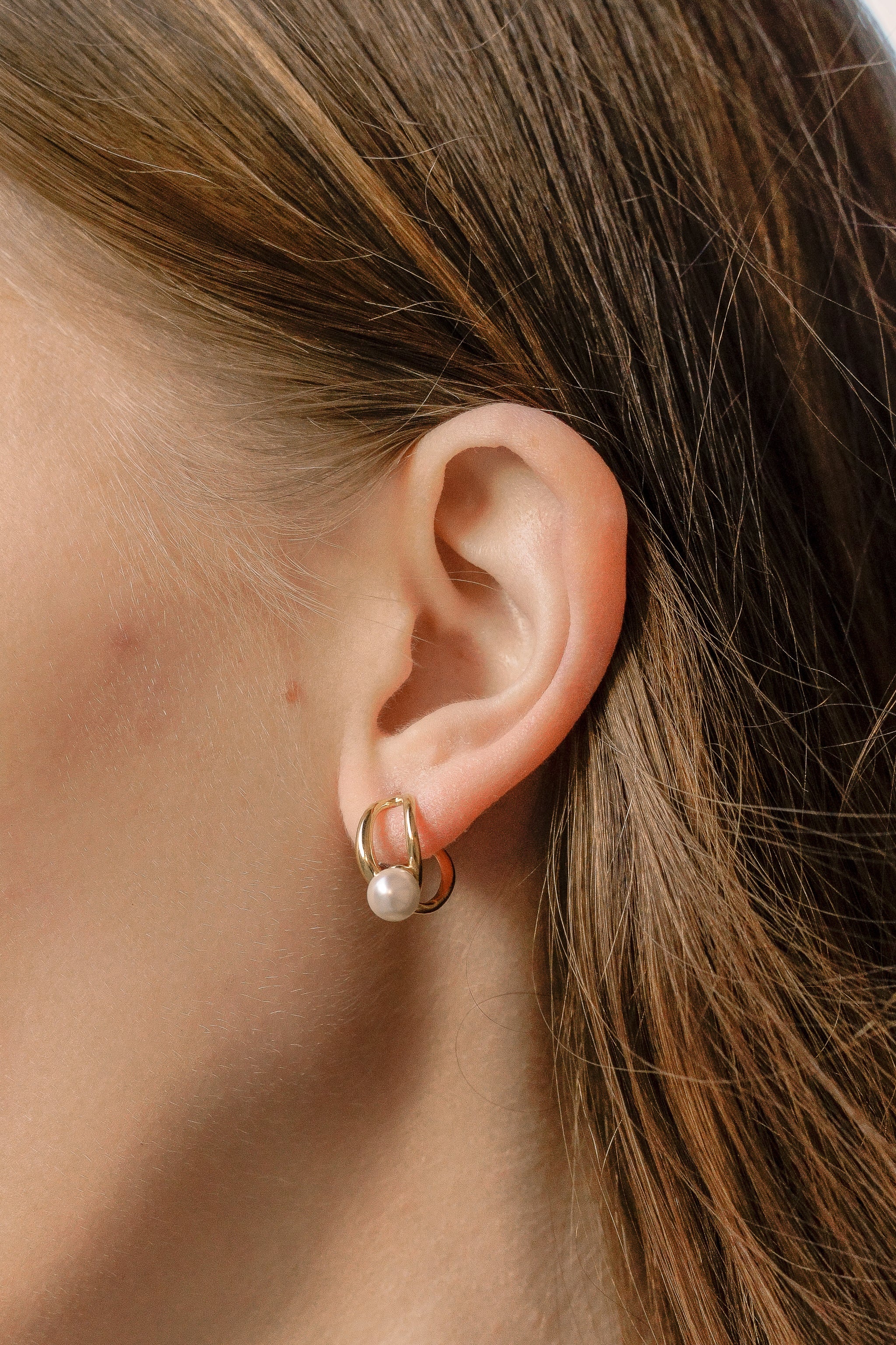 Mazie auskari - Loop by Como Jewelry, ar 14k zelta pārklājumu un pērles imitāciju. Brūni mati daļēji ierāmē seju, un gluda āda izceļ eleganto auskaru.