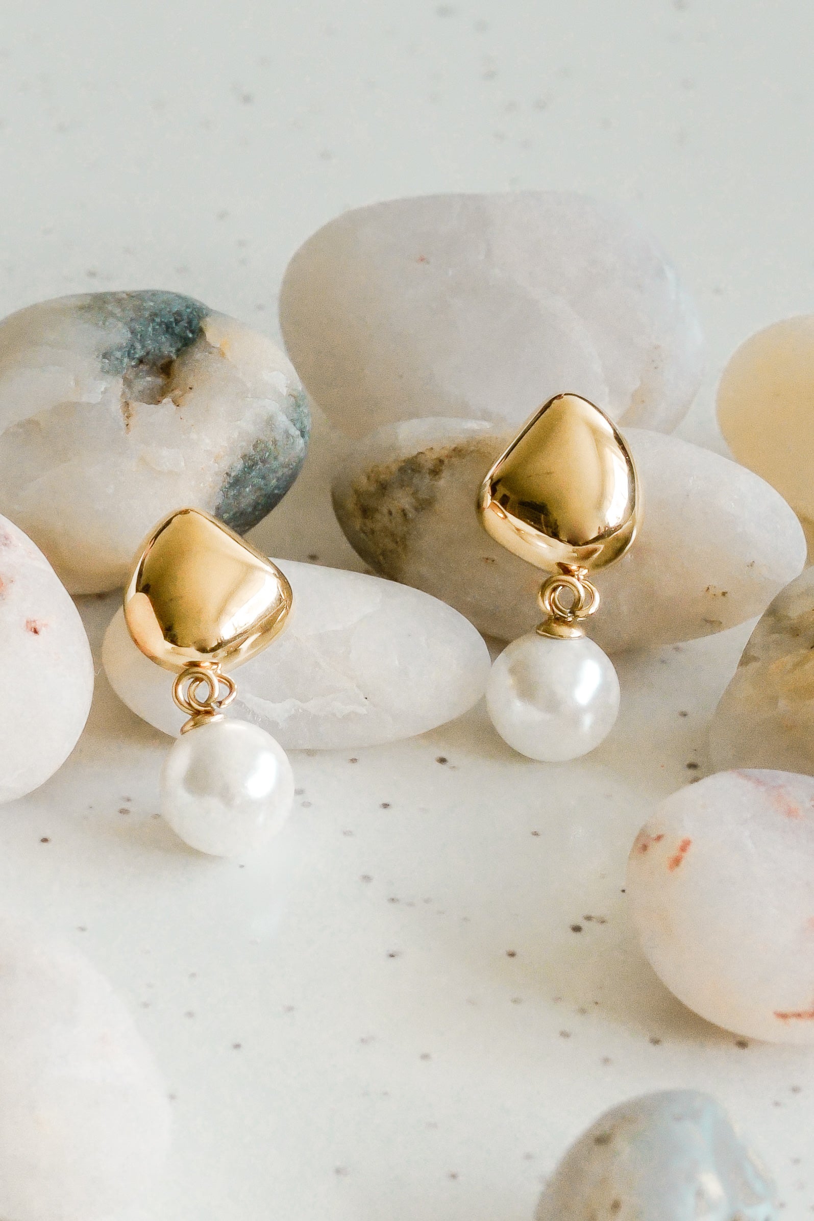 Mazie auskari - Rīta rasa no Como Jewelry ar zelta akcentiem ir izvietoti uz gludiem, gaišiem akmeņiem ar smalkiem plankumiem un dabiskām faktūrām fonā.