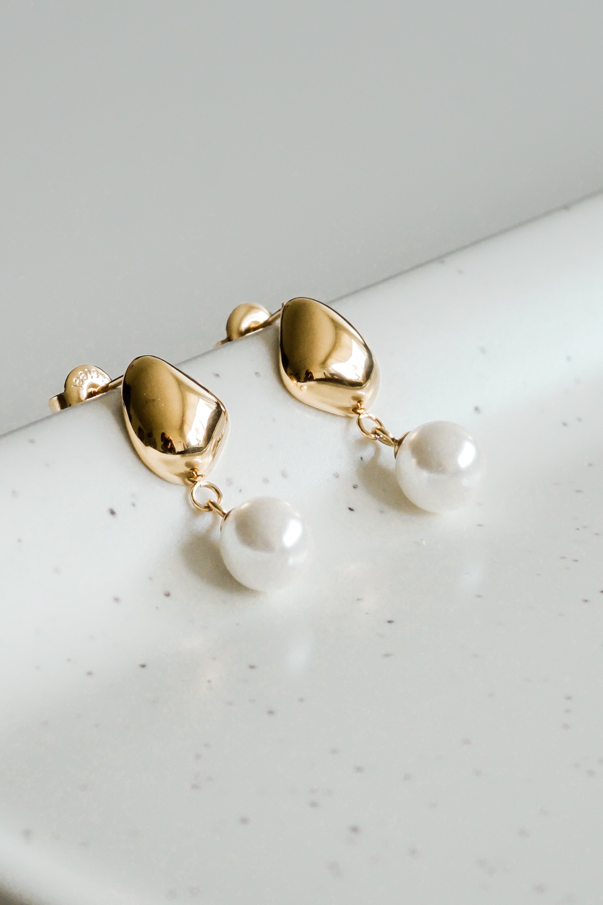 Mazie auskari - Rīta rasa no Como Jewelry ir hipoalerģiski zelta auskari ar gludām, noapaļotām virsmām un baltām pērlēm, kas izvietotas uz baltas raibas keramikas virsmas.