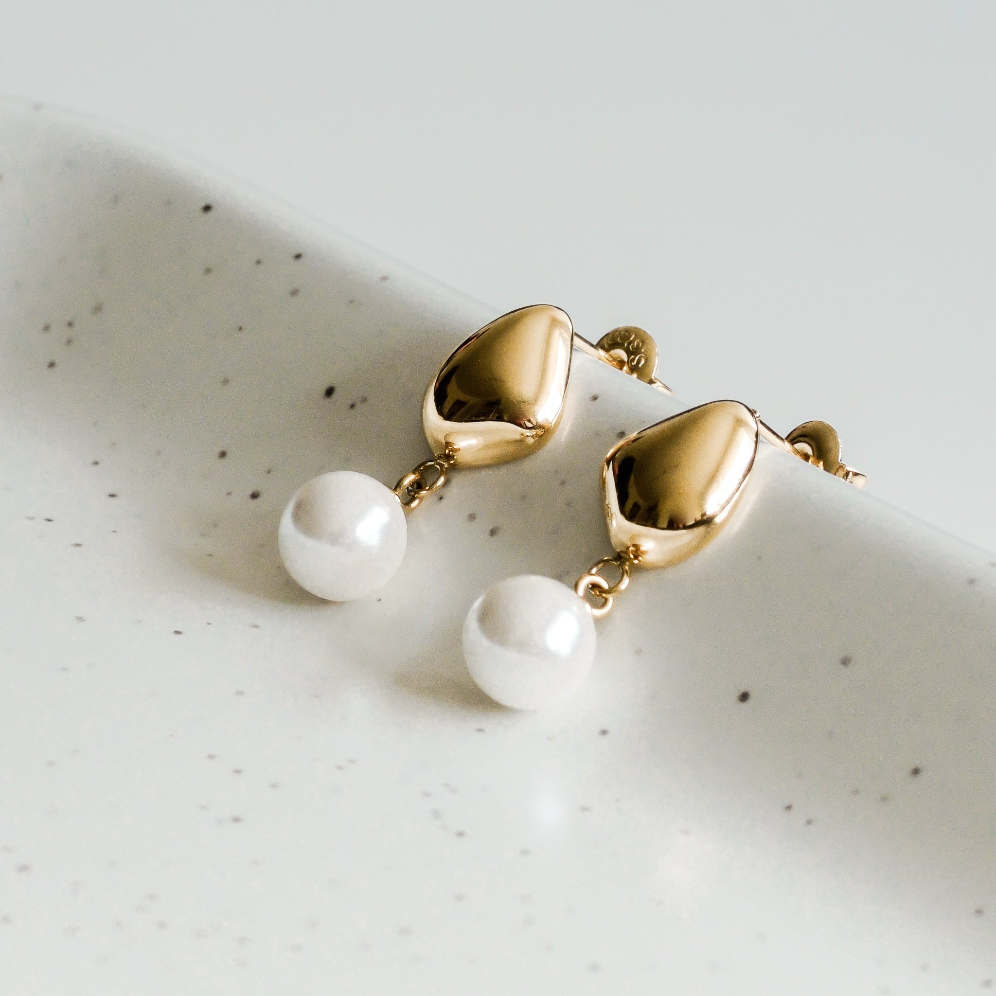 Mazie auskari - Rīta rasa by Como Jewelry ir ar asaru formas virsotnēm, zem kurām karājas baltas apaļas pērlītes, kas izvietotas uz baltas, plankumainas virsmas - eleganti auskari, kas piešķir izsmalcinātību jebkuram tērpam.