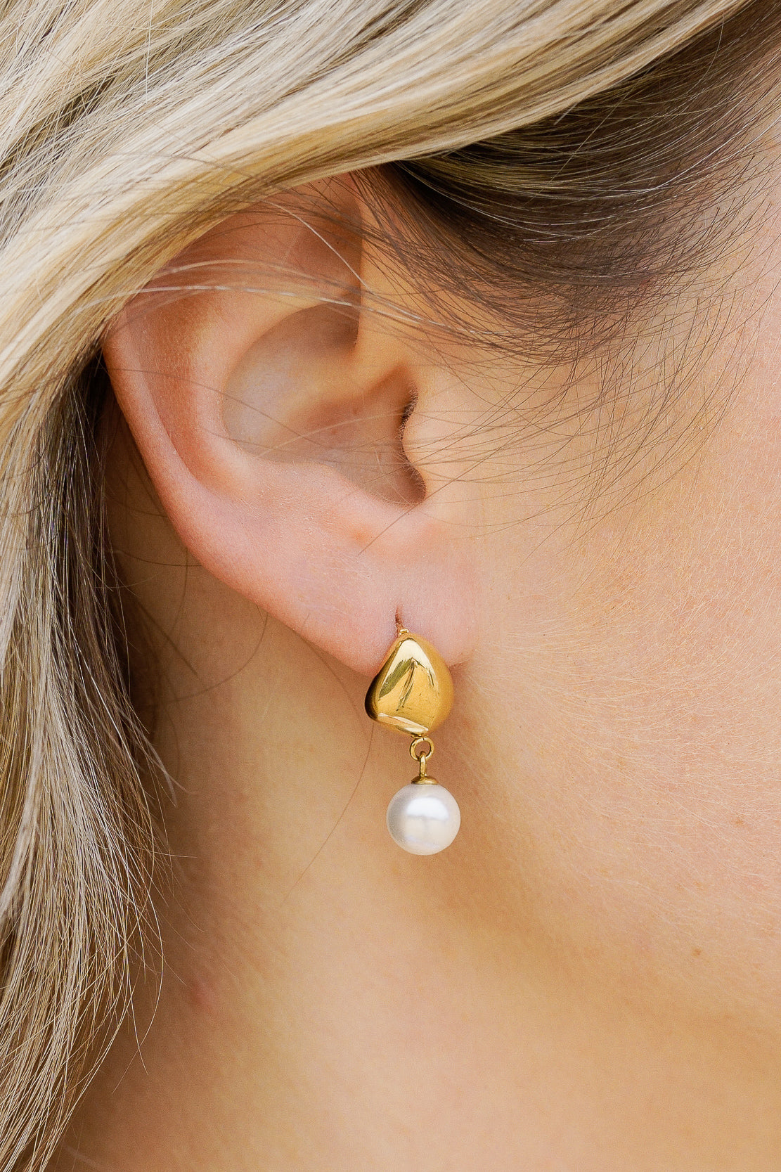 Tuvplāns sievietes ausij ar gaišu ādu un gaišiem matiem, kurai ir Mazie auskari - Rīta rasa no Como Jewelry, ar hipoalerģisku zeltu un piekarināmu baltu pērli.