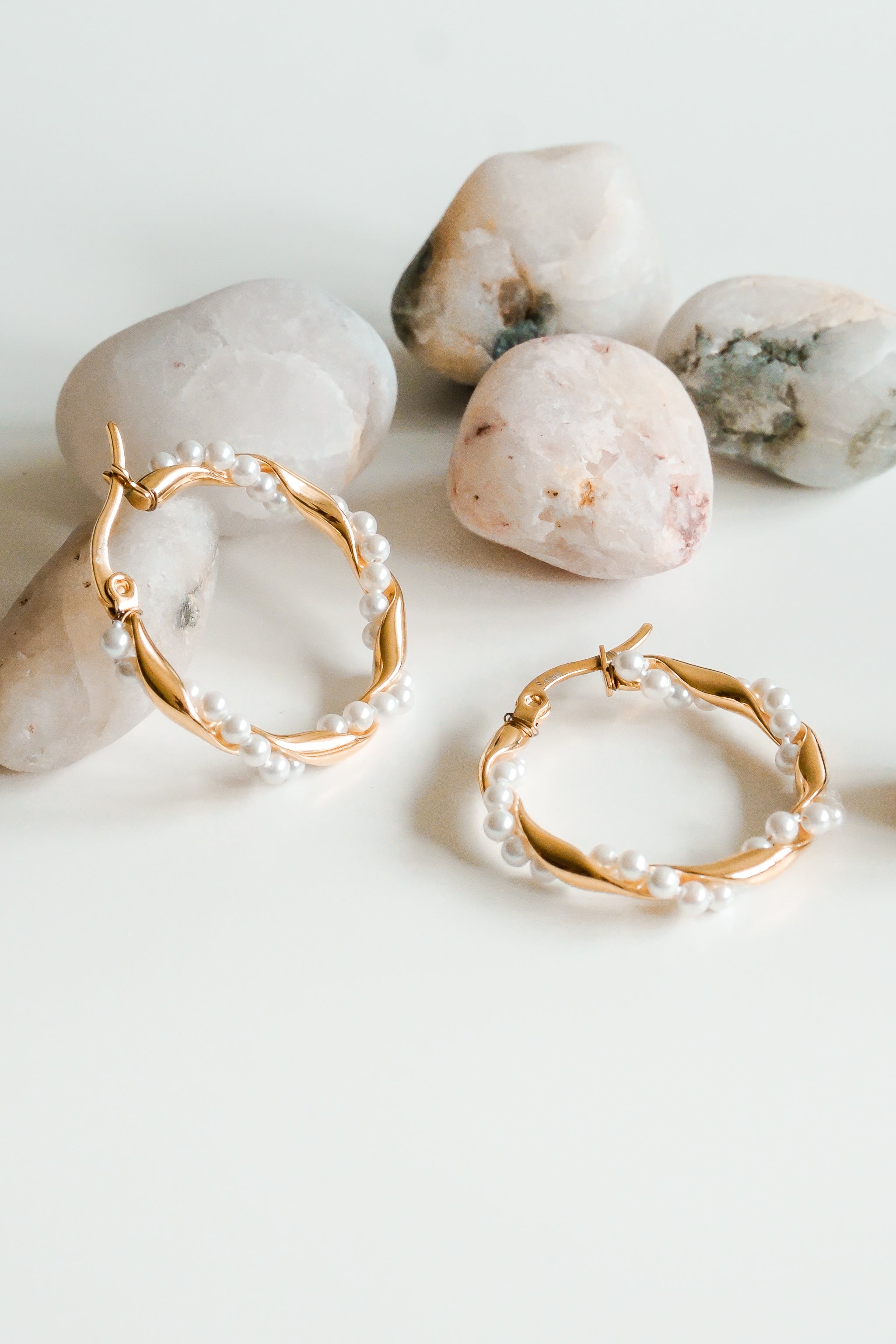 Como Jewelry Mazie auskari - Pērļu vainags ir ar mazām baltām pērlītēm ar zelta pārklājumu, kas attēlotas uz baltas virsmas blakus gludiem, gaišiem akmentiņiem.