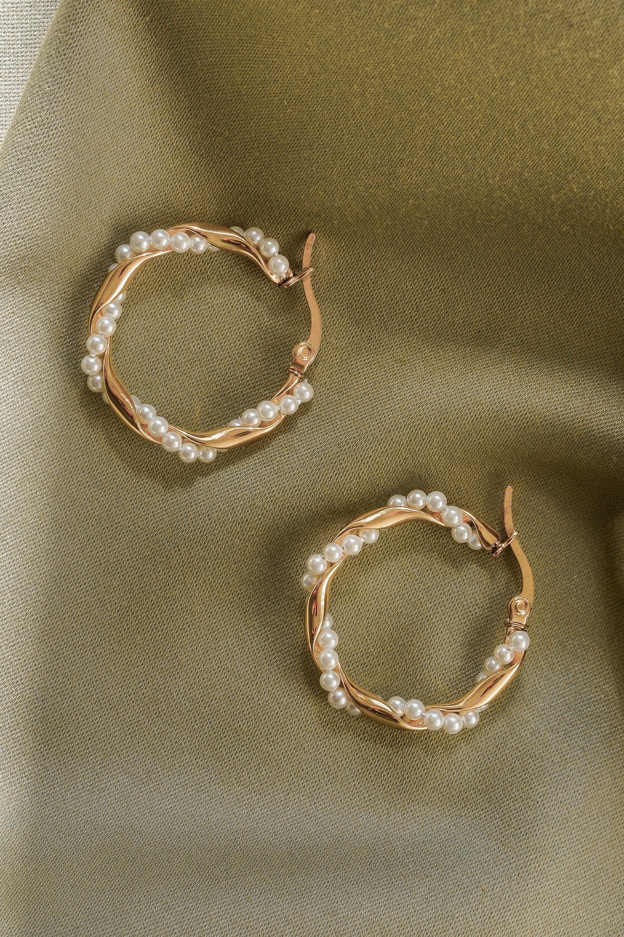 Como Jewelry Mazie auskari - Pērļu vainagi, rotāti ar mazām baltām pērlītēm, ir izvietoti uz fakturēta gaiši zelta auduma fona.