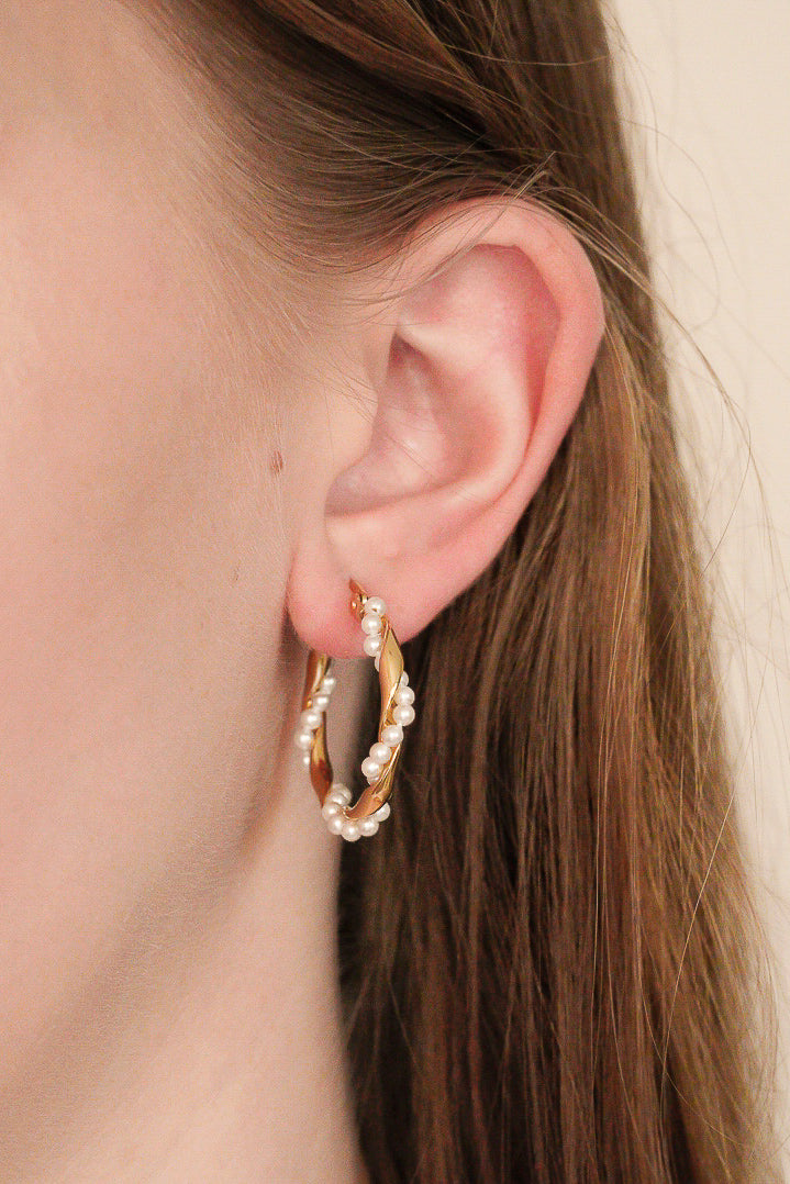 Tuvuplāns sievietes ausij ar Como Jewelry "Mazie auskari - Pērļu vainags"; viņas gaiši brūnie mati ir nolaisti atpakaļ, izceļot apzeltīto cilpu, ko rotā maigas baltas pērlītes.